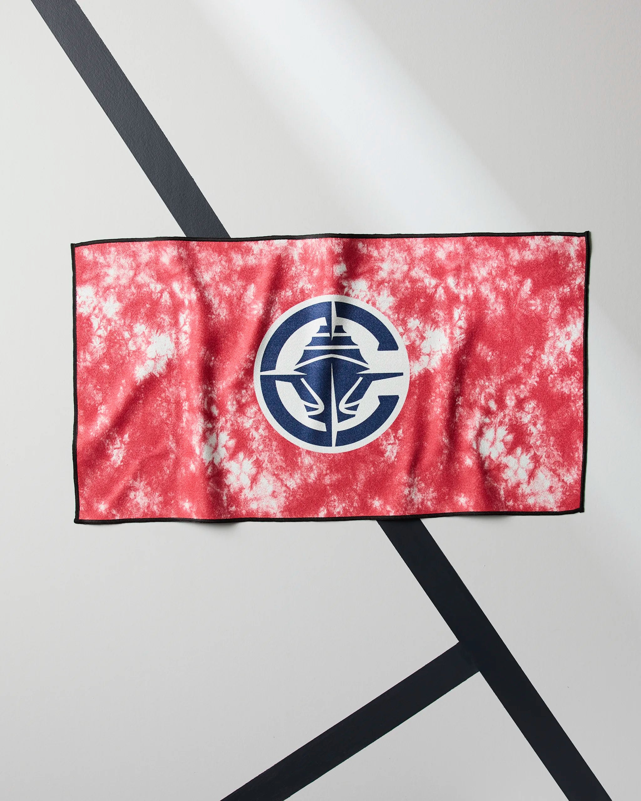 The Geometry Clippers Flash is a red and white tie-dye ultra-absorbent workout towel with a black border and a large blue and white circular logo of stylized hands around a lighthouse, shown flat on a diagonally striped surface.