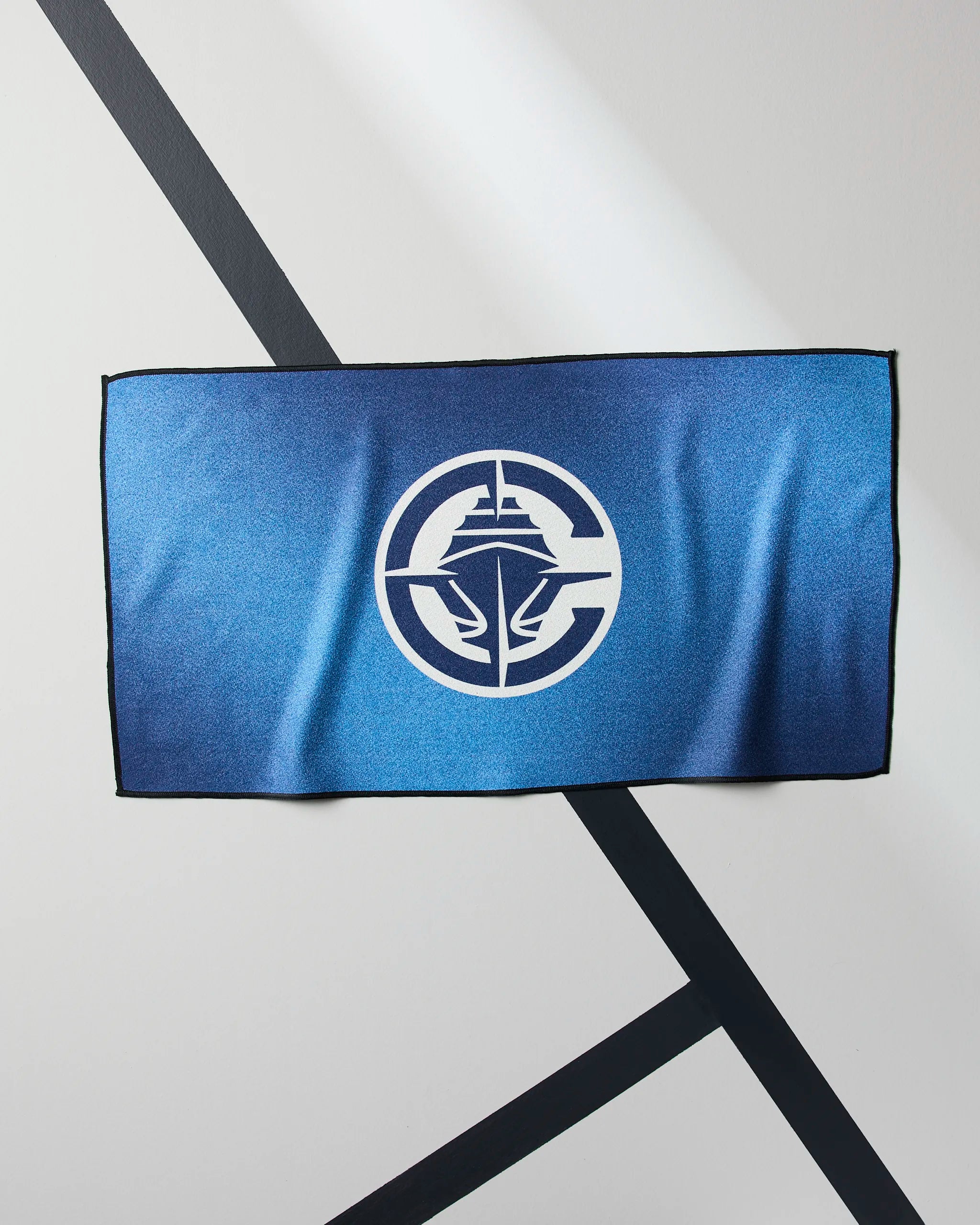 A blue rectangular Clippers Fade towel by Geometry, featuring a white circular logo with a stylized ship and waves, hangs on a light-colored wall with diagonal black lines.