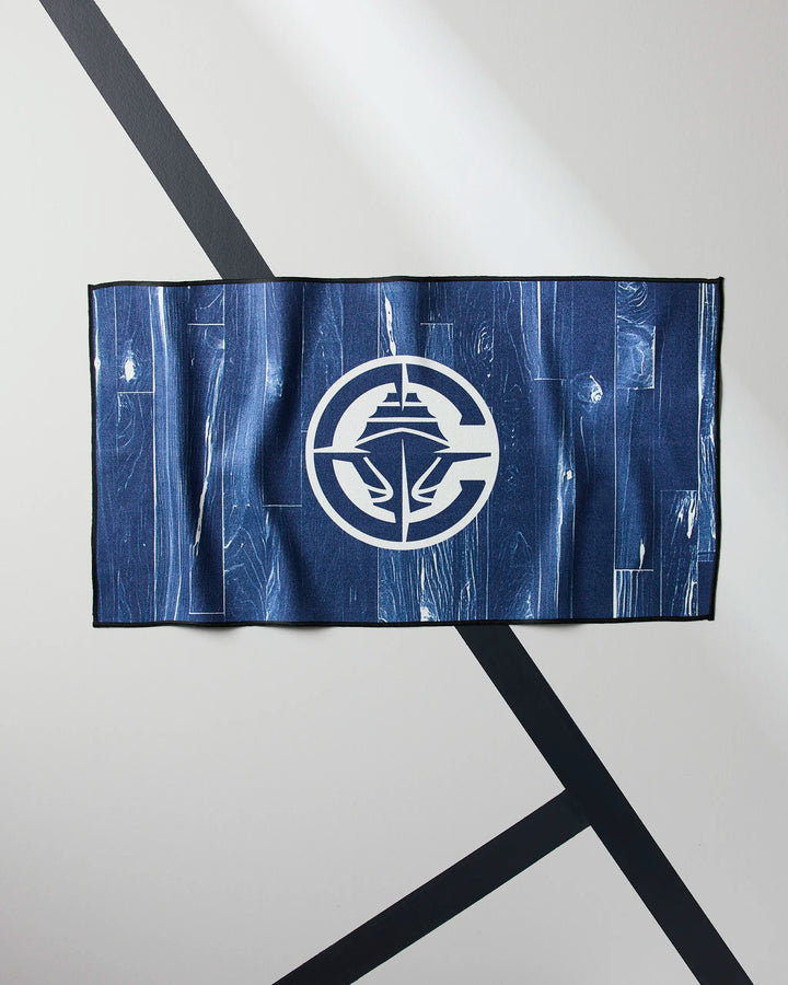 The Clippers Zone gym towel from NBALAB x Geometry is blue, rectangular, and features a white abstract circular logo in the center. It displays a unique wood grain pattern and is shown on a gray surface with black intersecting diagonal lines.