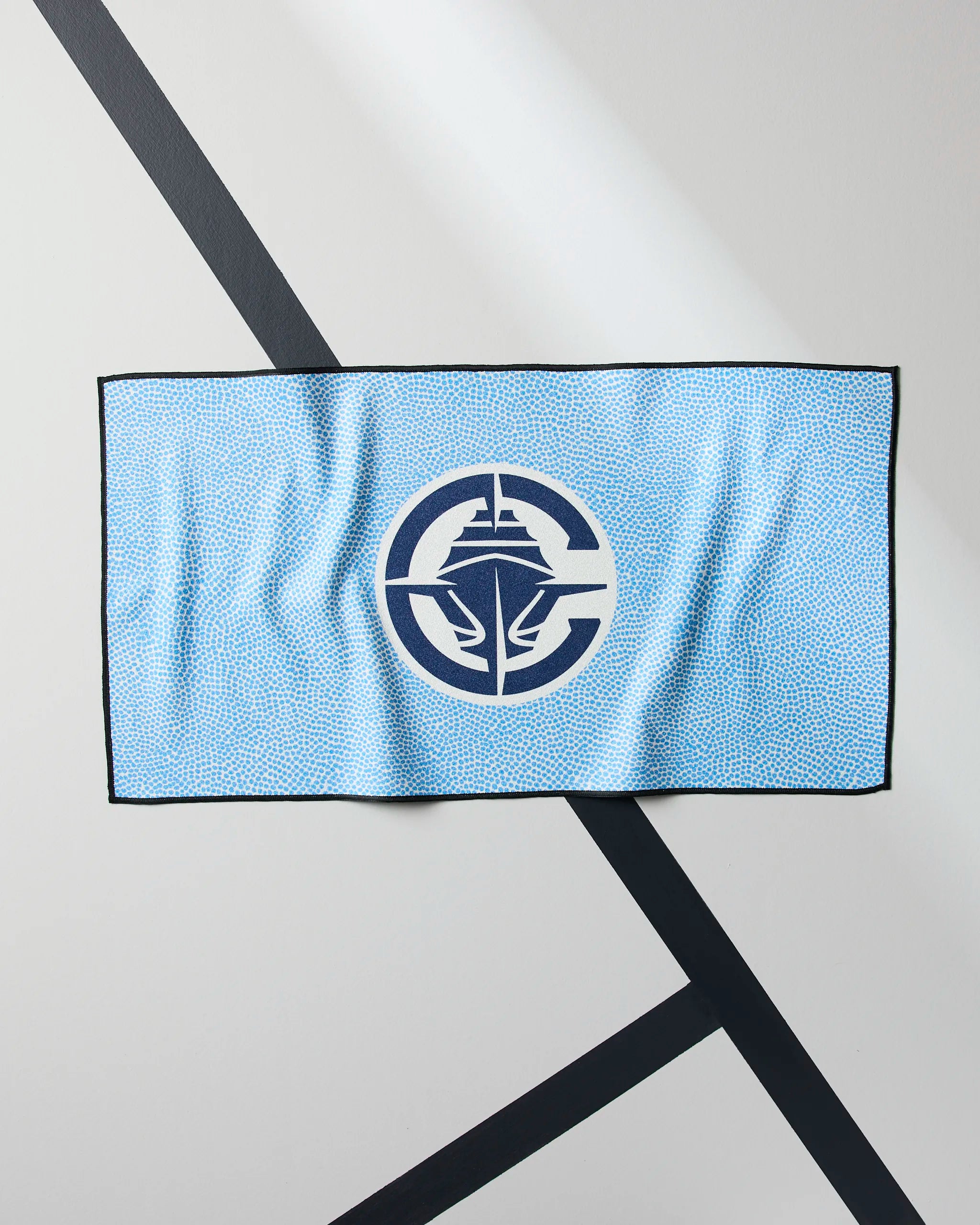 A light blue Clippers Grip gym towel by Geometry, featuring a quick-drying design, black border, and a large navy and white ship logo inside a circle at the center, rests on a white surface with intersecting black lines.