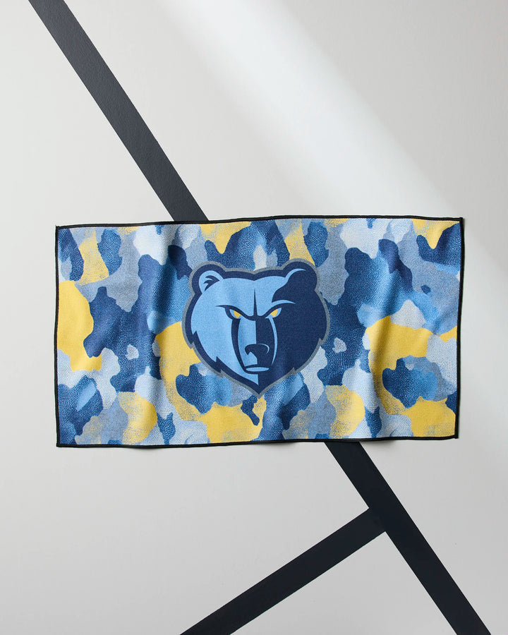 The Grizzlies Camo gym towel by NBALAB x Geometry has a blue and yellow camouflage design with the Memphis Grizzlies bear logo in the center, displayed on a white wall with black diagonal lines.