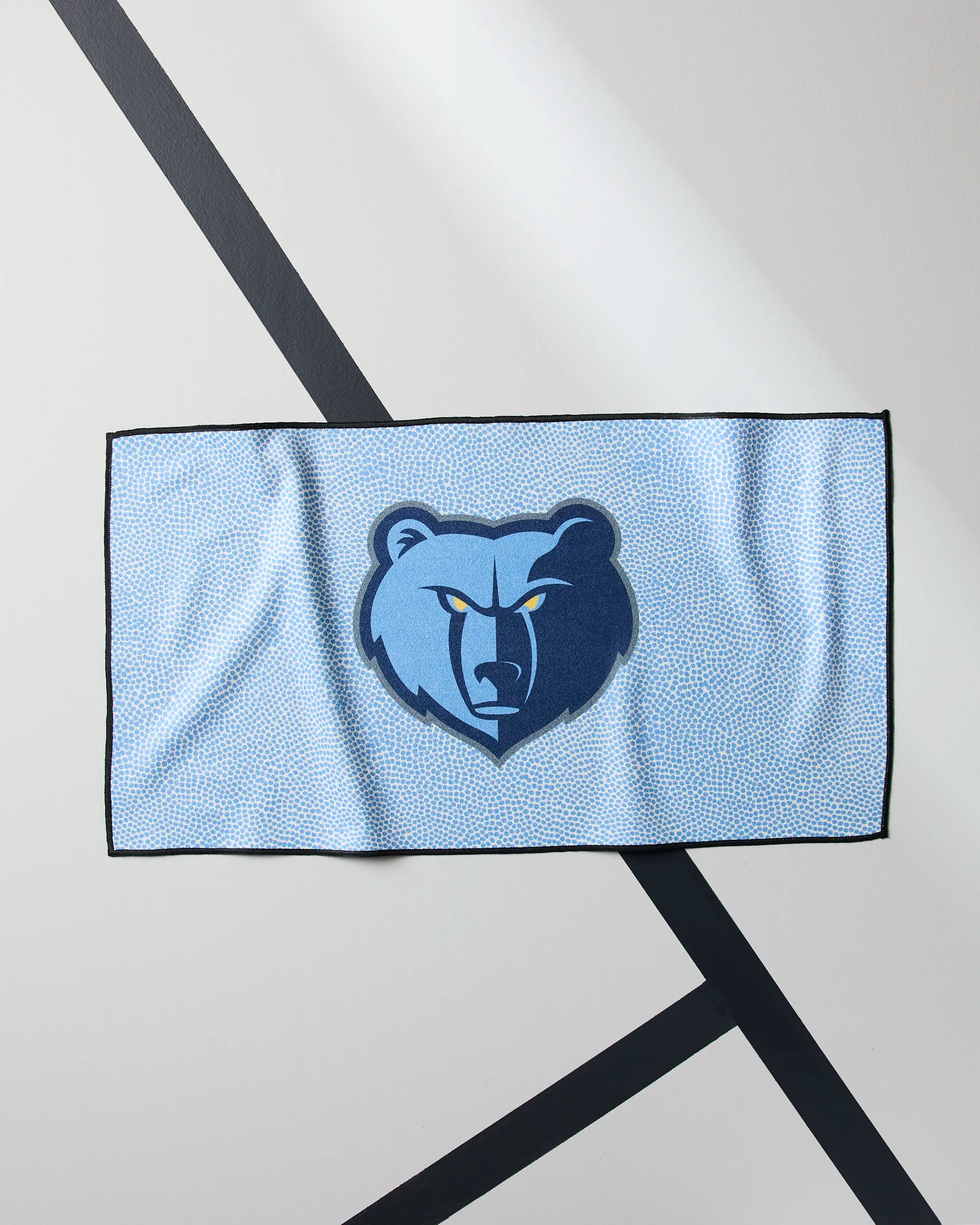 The Grizzlies Grip by Geometry is a light blue, quick-drying gym towel with the Memphis Grizzlies bear logo in dark and light blue on a white background with diagonal black lines—ideal for NBA fans during workouts.