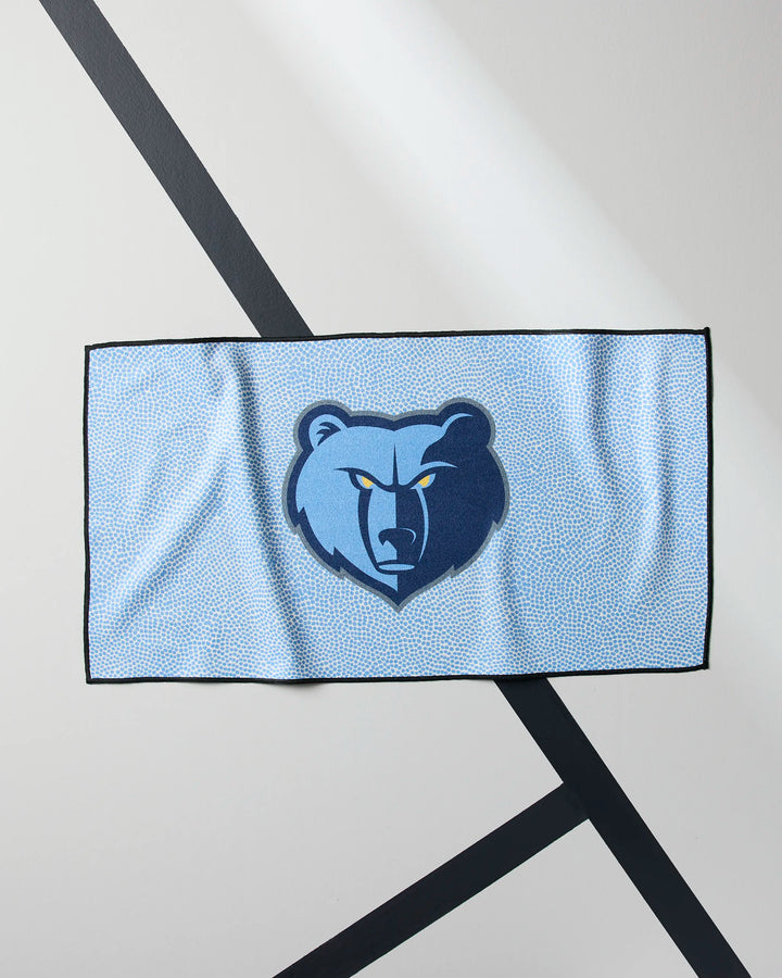 The NBALAB x Geometry Grizzlies Grip is a light blue, quick-drying gym towel with a dark and light blue stylized bear head logo, displayed flat on a light gray surface with intersecting black diagonal lines.