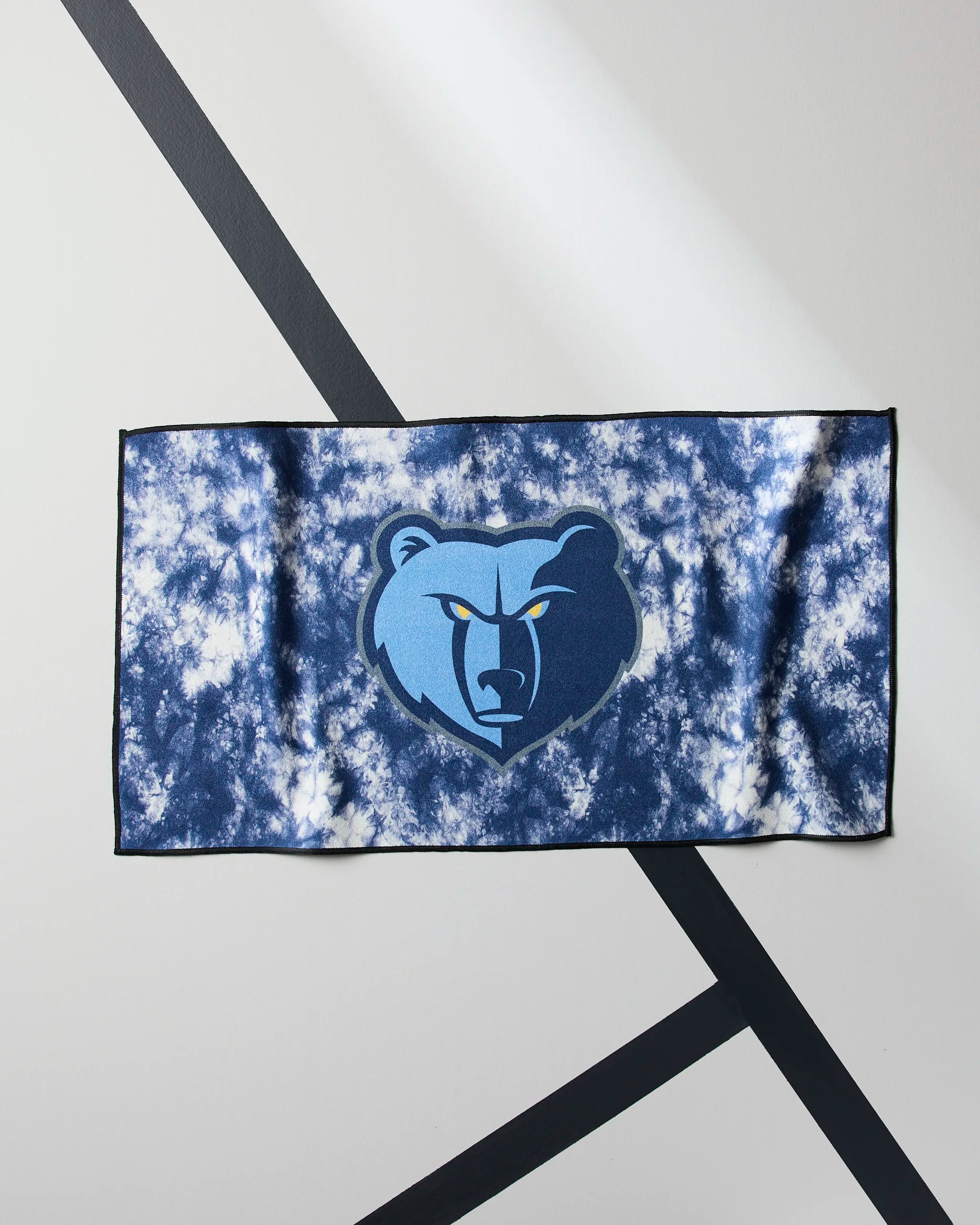 The Geometry Grizzlies Flash NBA fitness towel, featuring a blue and white tie-dye design with the Memphis Grizzlies bear logo, is laid flat on a gray surface with diagonal black lines.