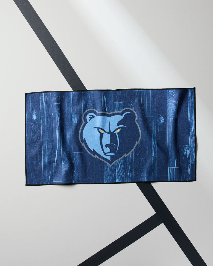 The Grizzlies Zone fitness towel by NBALAB x Geometry is a blue, rectangular NBA towel with the Memphis Grizzlies bear logo centered, shown on a light surface with dark diagonal lines in the background.