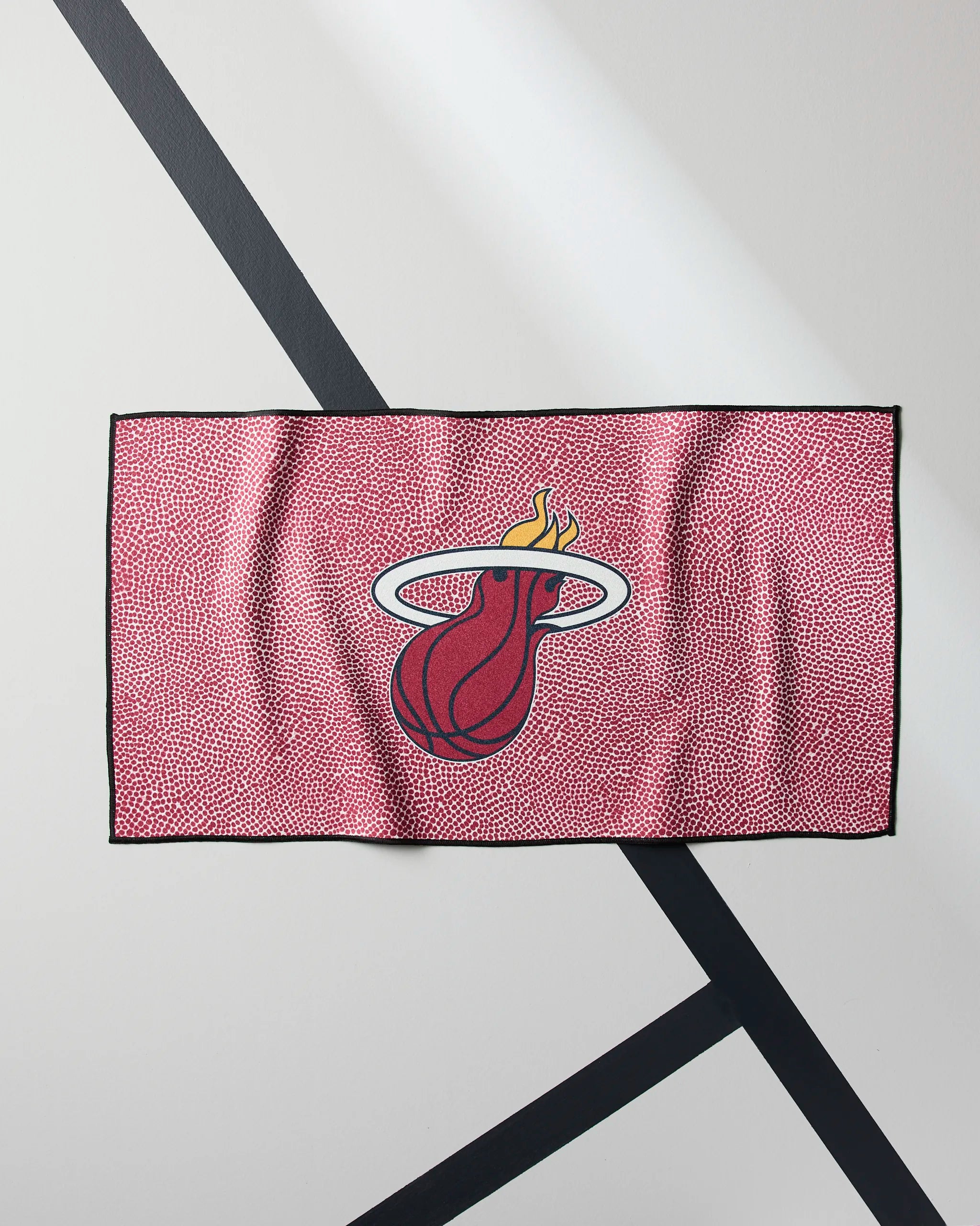 A red Heat Grip fitness towel by Geometry, featuring the Miami Heat logo, hangs against a white wall with diagonal black lines.