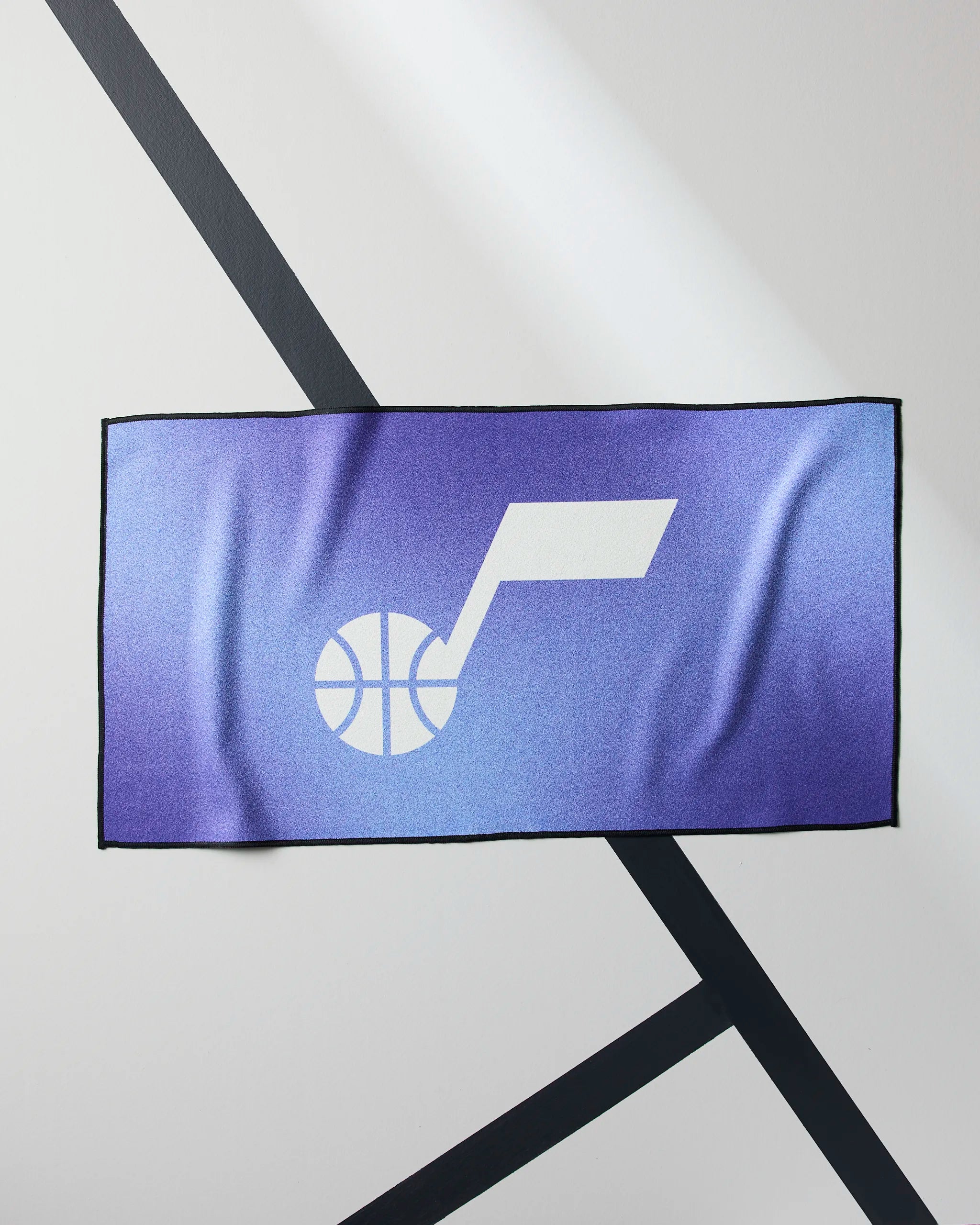 The Geometry Jazz Fade towel is a rectangular purple NBA fitness towel featuring a white basketball and musical note logo centered on its front, displayed on a light background with intersecting black lines.