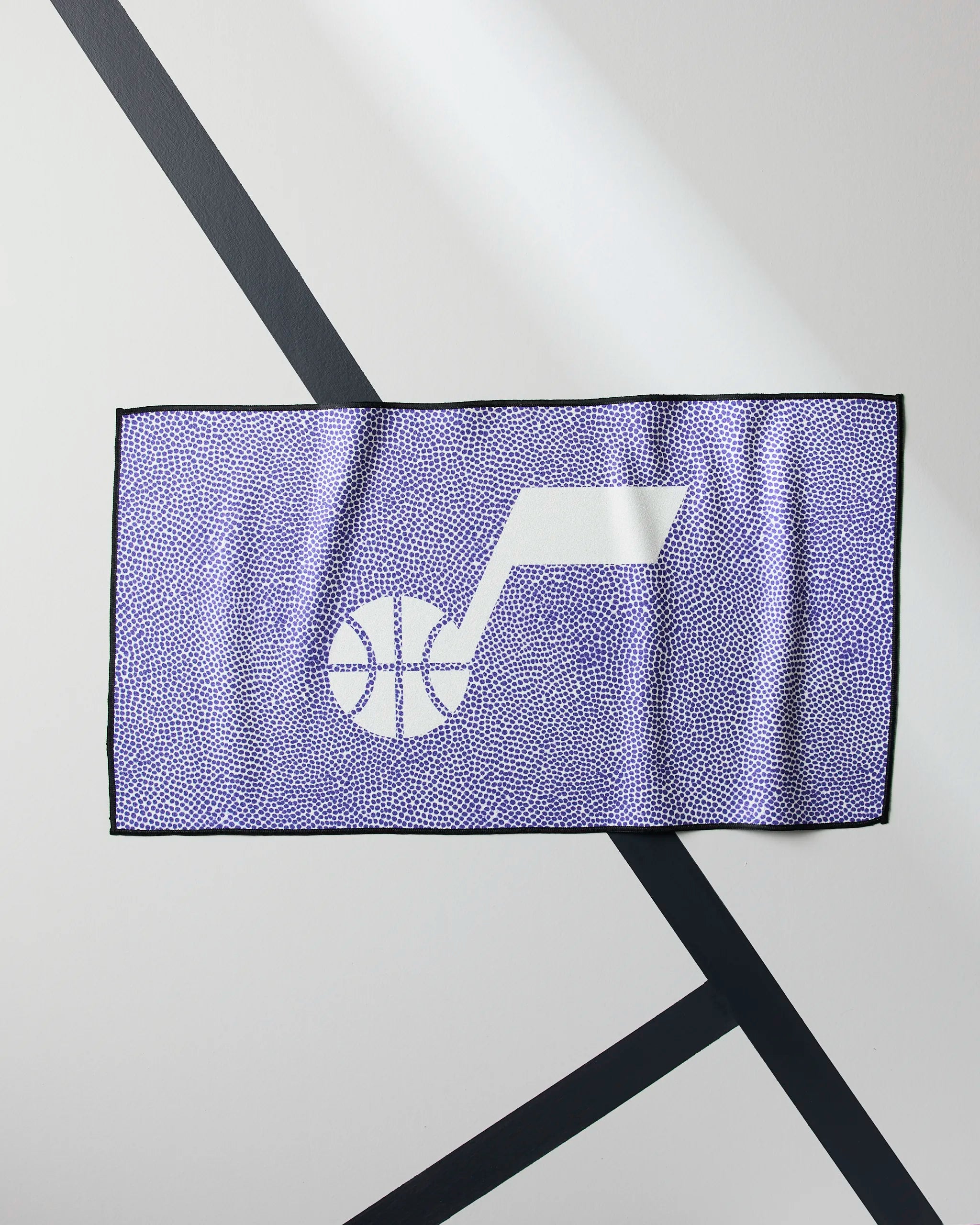 The Geometry Jazz Grip is a purple quick-drying workout towel with a white Utah Jazz basketball logo, shown lying flat on a white surface with black diagonal lines beneath it.