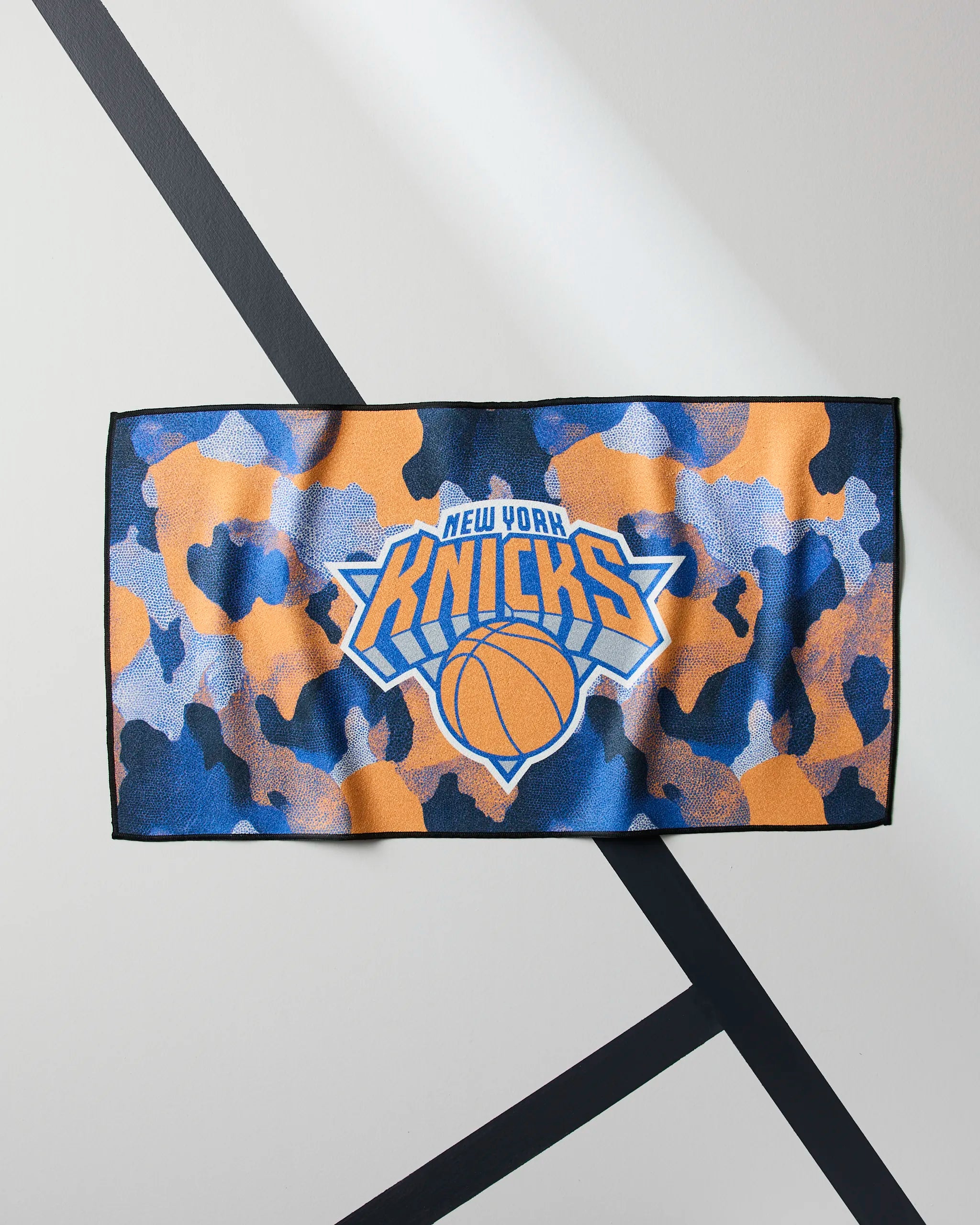 The Geometry Knicks Camo towel, featuring a blue, orange, and black Knicks camo pattern with the team's logo, is displayed flat on a white surface with intersecting black lines.
