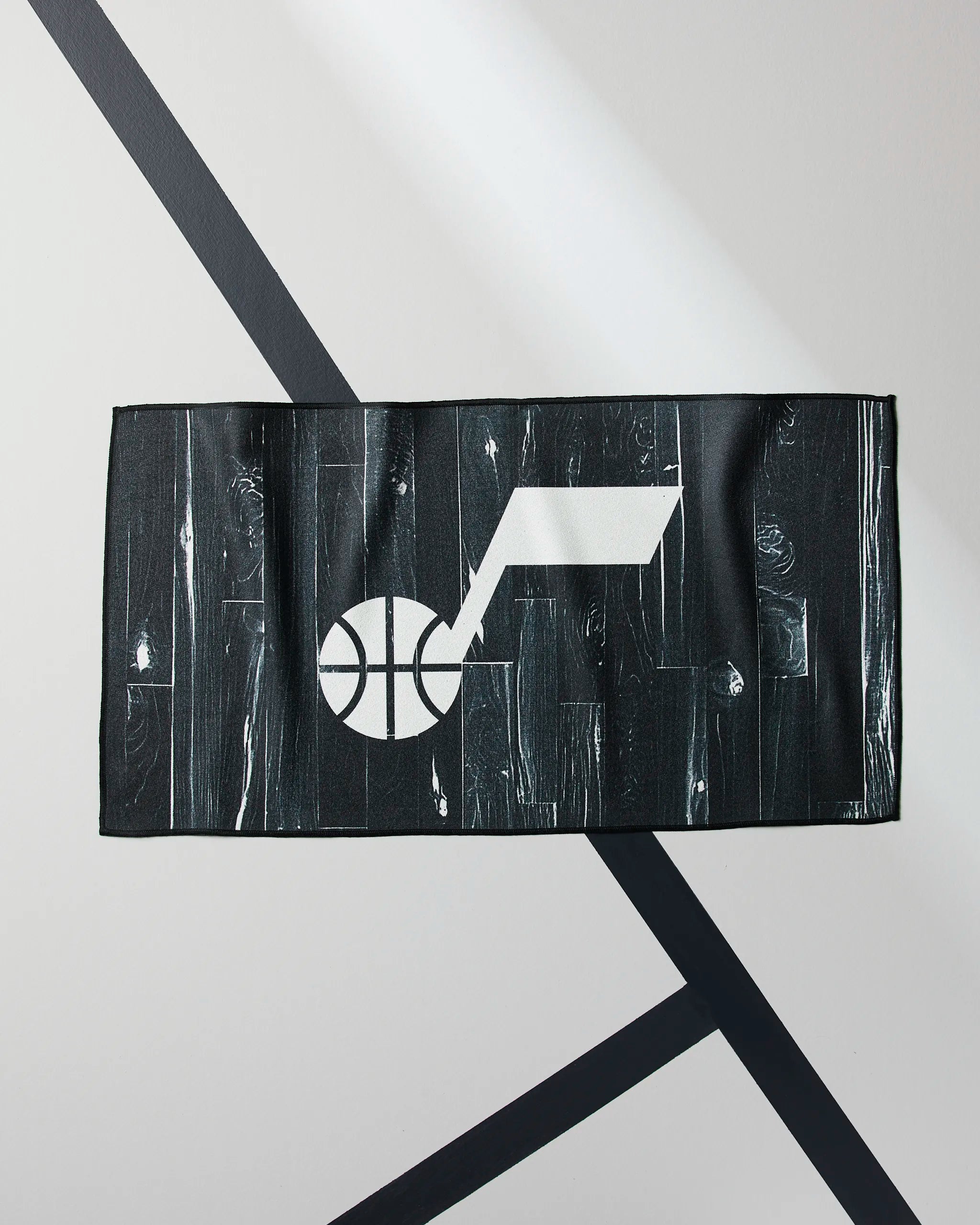 The Geometry Jazz Zone is a rectangular black NBA fitness towel featuring a white stylized basketball and note graphic, displayed on a light gray surface with diagonal black lines.