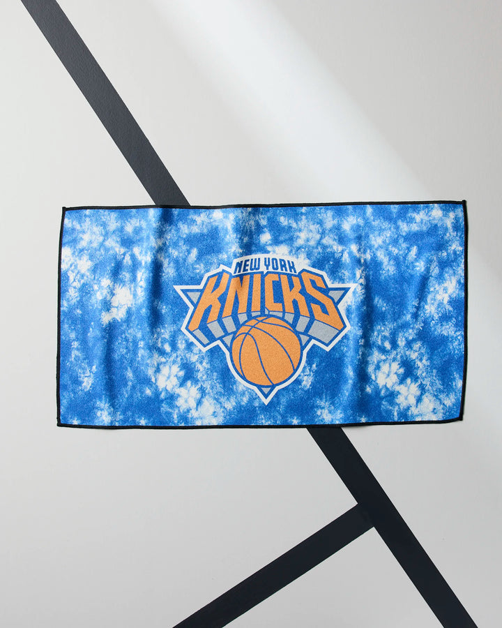The Knicks Flash by NBALAB x Geometry is a blue and white tie-dye NBA fitness towel with the New York Knicks logo centered. Laid flat on a white surface with black diagonal lines, it's perfect as a quick-drying gym towel for fans and athletes.