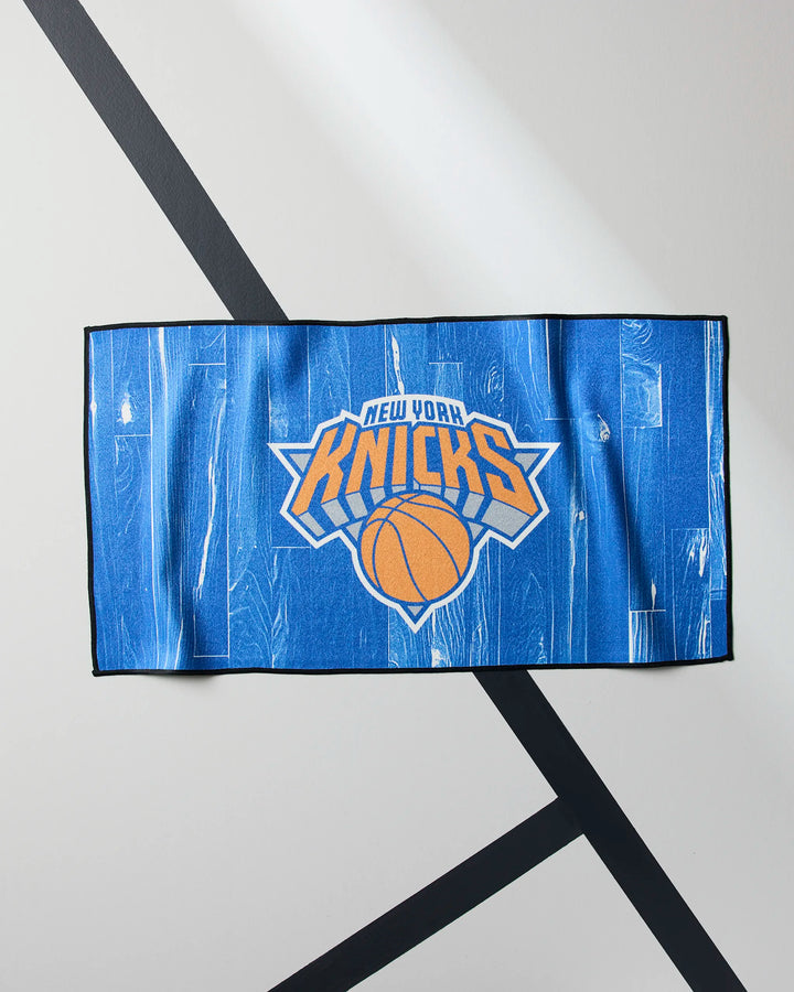 The Knicks Zone towel by NBALAB x Geometry, featuring the New York Knicks logo with an orange basketball and white lettering on blue, is laid flat on a light surface with black diagonal lines.