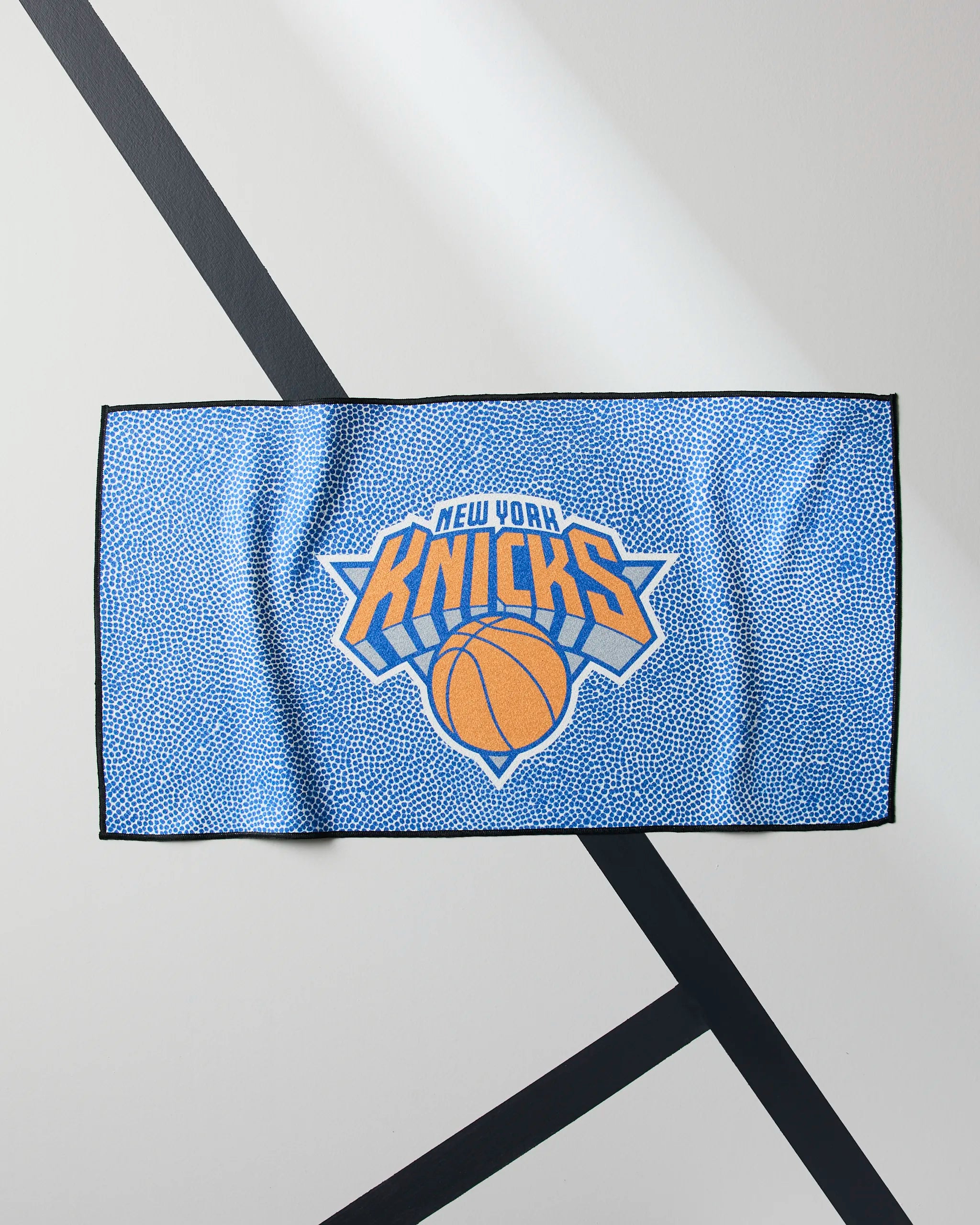 The Geometry Knicks Grip is a blue, quick-drying workout towel with a dotted pattern and New York Knicks logo lying flat on a white background with black diagonal lines.
