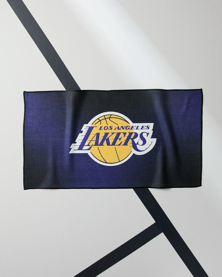 The Lakers Fade by NBALAB x Geometry is a rectangular fitness towel with the Los Angeles Lakers logo—yellow basketball and purple text on a dark background—laid flat with diagonal black lines. Quick-drying and perfect for any workout.