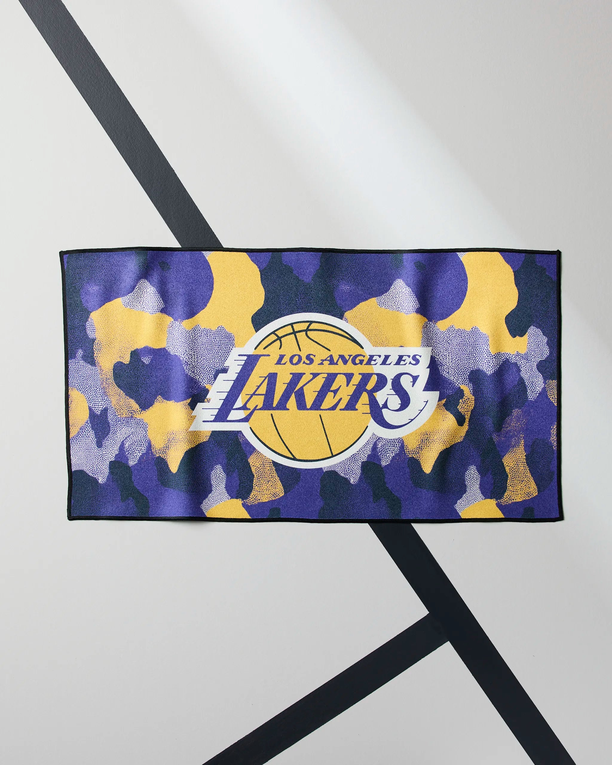The Geometry Lakers Camo towel is rectangular, quick-drying, and features a yellow, purple, and black camouflage design with the Los Angeles Lakers logo on a light gray background with dark diagonal lines.