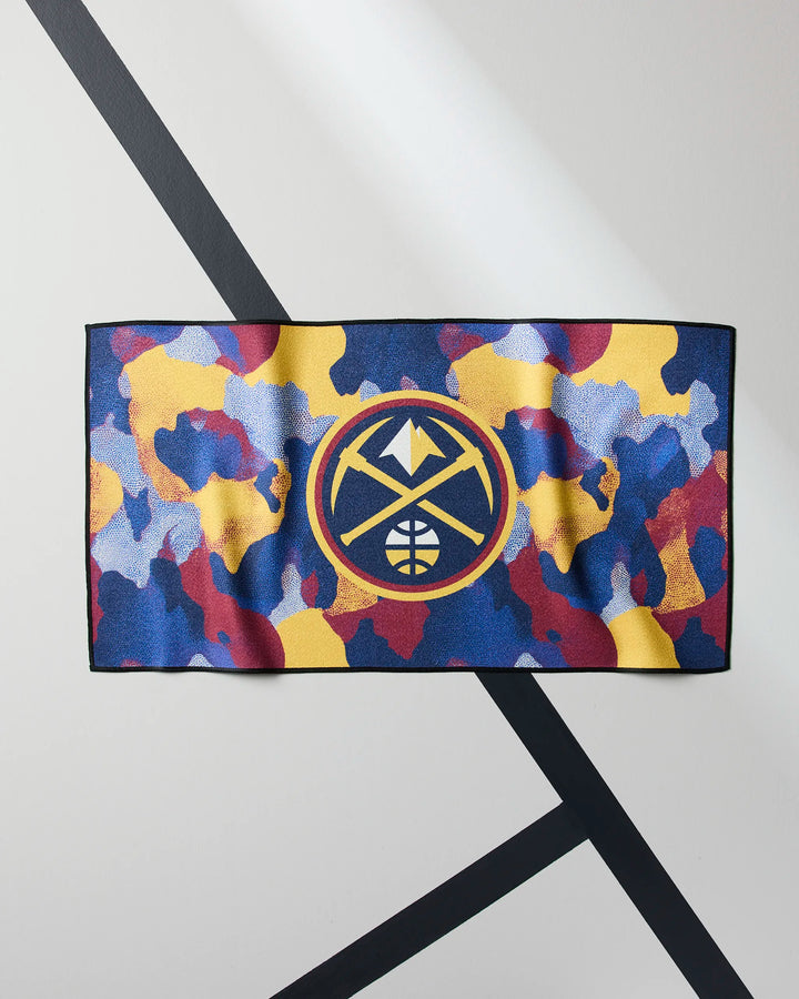 The NBALAB x Geometry "Nuggets Camo" fitness towel features a blue, yellow, red, and purple camouflage design with the Denver Nuggets logo at the center, displayed against a light wall with black diagonal lines.