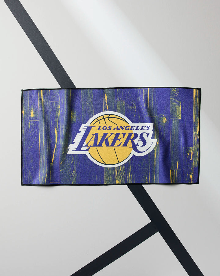 The Lakers Zone fitness towel by NBALAB x Geometry, featuring the Los Angeles Lakers logo on a purple and gold abstract background, is displayed on a white surface with black diagonal lines.
