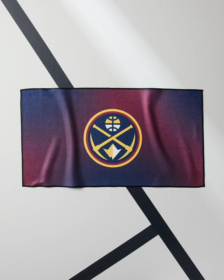 The NBALAB x Geometry Nuggets Fade gym towel, featuring the Denver Nuggets gold and blue logo, lies flat on a diagonally striped light and dark gray surface.