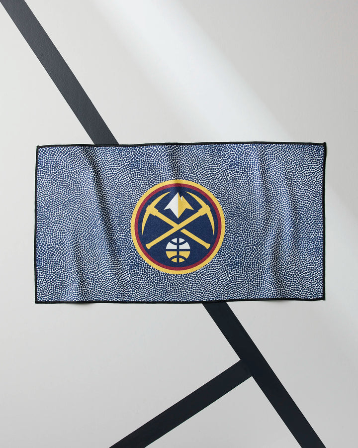 The Nuggets Grip by NBALAB x Geometry is a blue NBA fitness towel with the Denver Nuggets logo centered, made from Geoweave fabric and shown against a white wall with black diagonal lines.