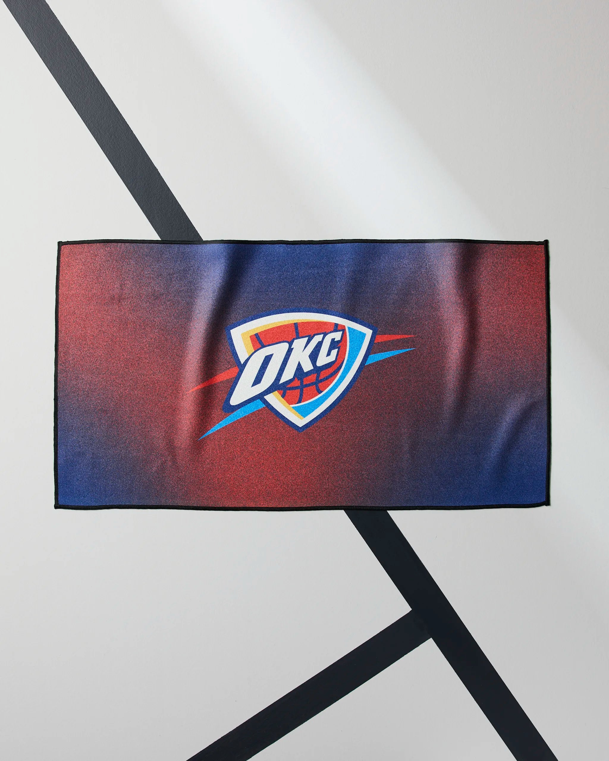 The Geometry OKC Fade towel is a rectangular NBA fitness towel showcasing the Oklahoma City Thunder logo over a gradient blue and red background, displayed on a white surface with black diagonal lines.