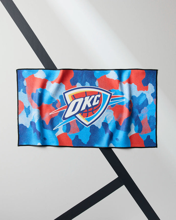 The OKC Camo towel by NBALAB x Geometry features a blue, red, and white camouflage pattern with the Oklahoma City Thunder logo at the center, shown on a light wall with diagonal black lines.