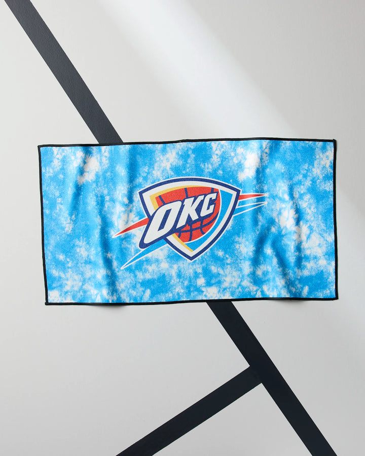The OKC Flash fitness towel by NBALAB x Geometry, boasting a blue and white tie-dye design with the Oklahoma City Thunder logo, hangs on a white wall accented by black diagonal lines.