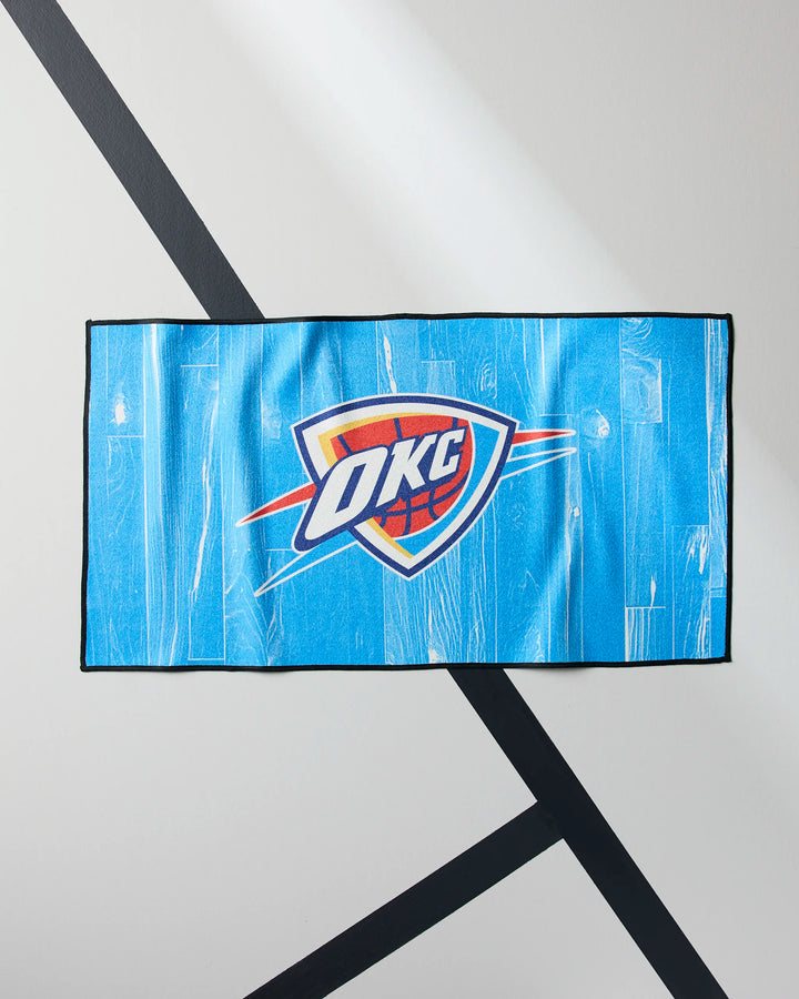 The blue OKC Zone fitness towel by NBALAB x Geometry, featuring the Oklahoma City Thunder logo, is displayed flat on a white and gray surface with diagonal black lines.