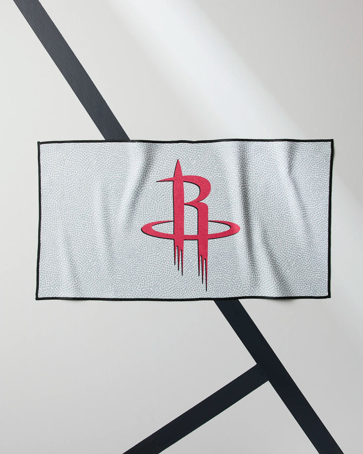 The Rockets Grip by NBALAB x Geometry is a sustainable gym towel with a black border and red Houston Rockets logo—ideal for NBA fans and fitness lovers. Displayed on a light surface with black diagonal lines.