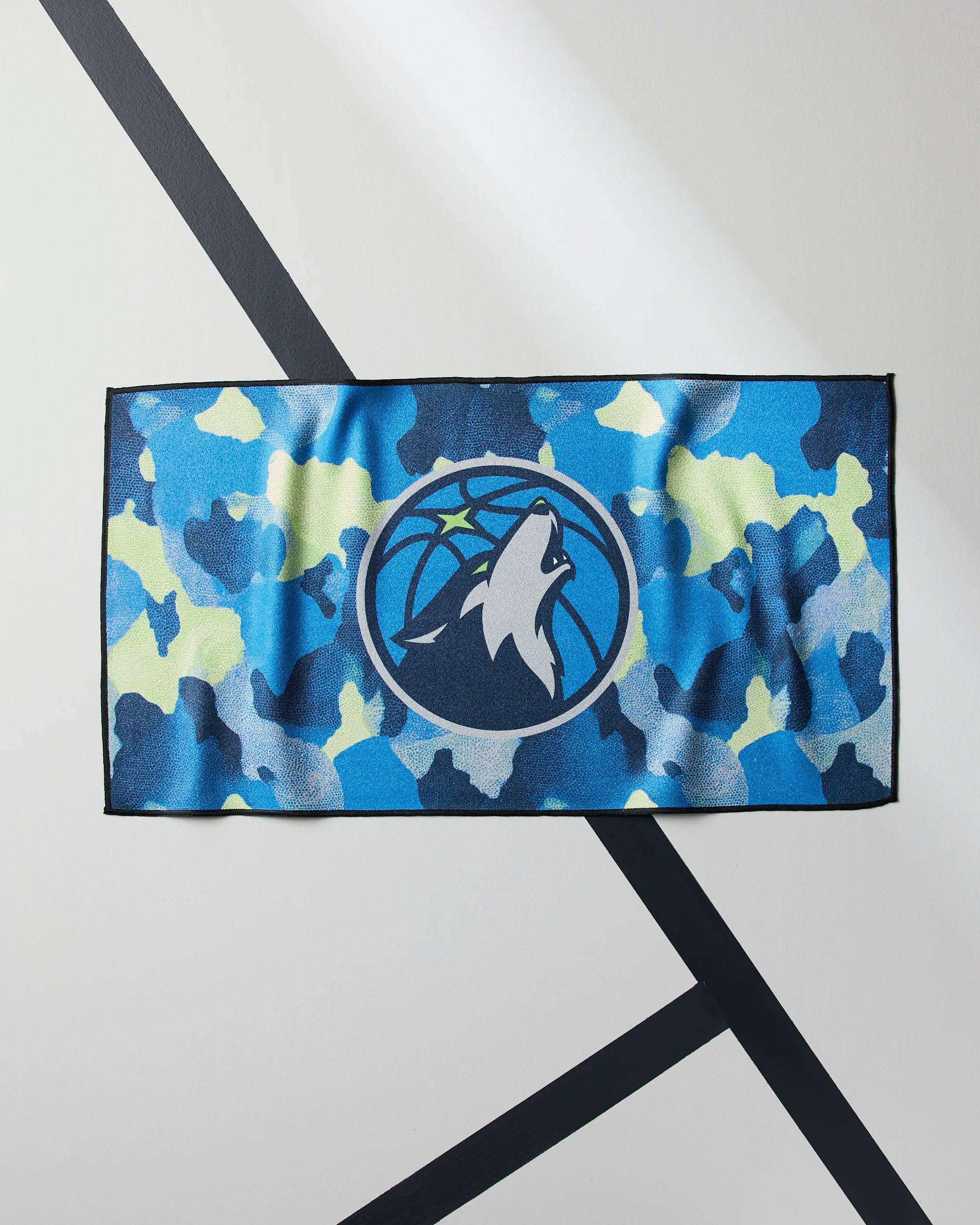 The Geometry Timberwolves Camo NBA fitness towel displays the Minnesota Timberwolves logo—a wolf howling before a basketball—on blue, green, and black camo and is laid flat on a white surface with black diagonal lines.