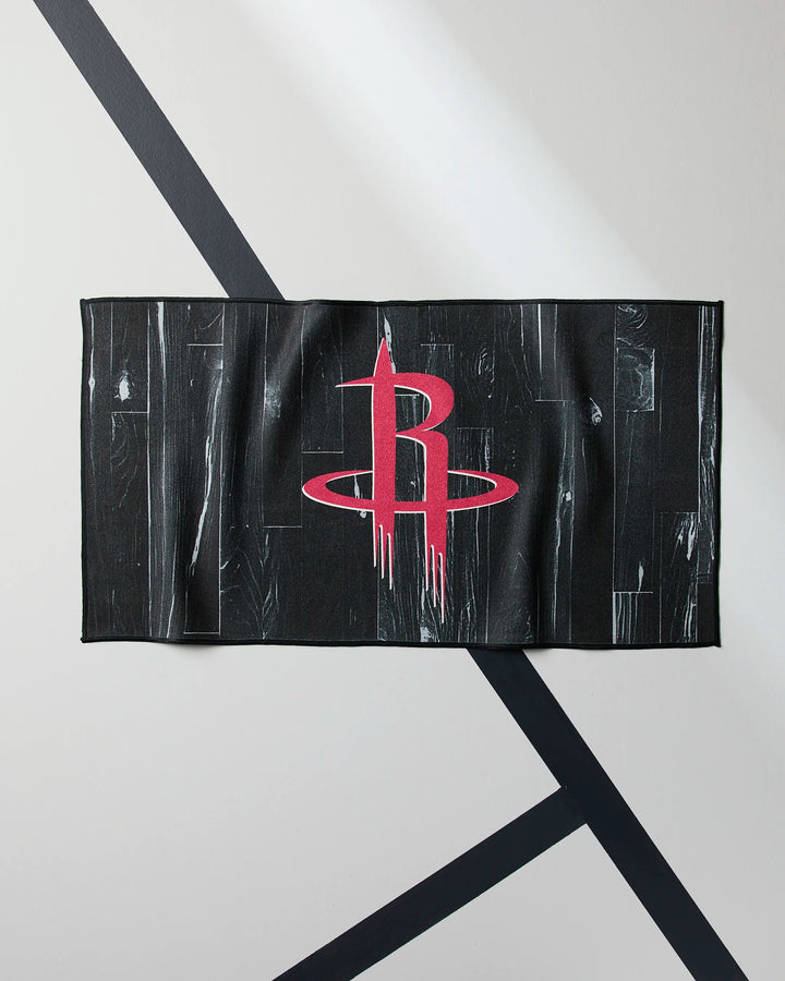 The Rockets Zone fitness towel by NBALAB x Geometry features a red Houston Rockets logo and wood grain pattern on black, displayed draped against a light wall with intersecting black diagonal lines.