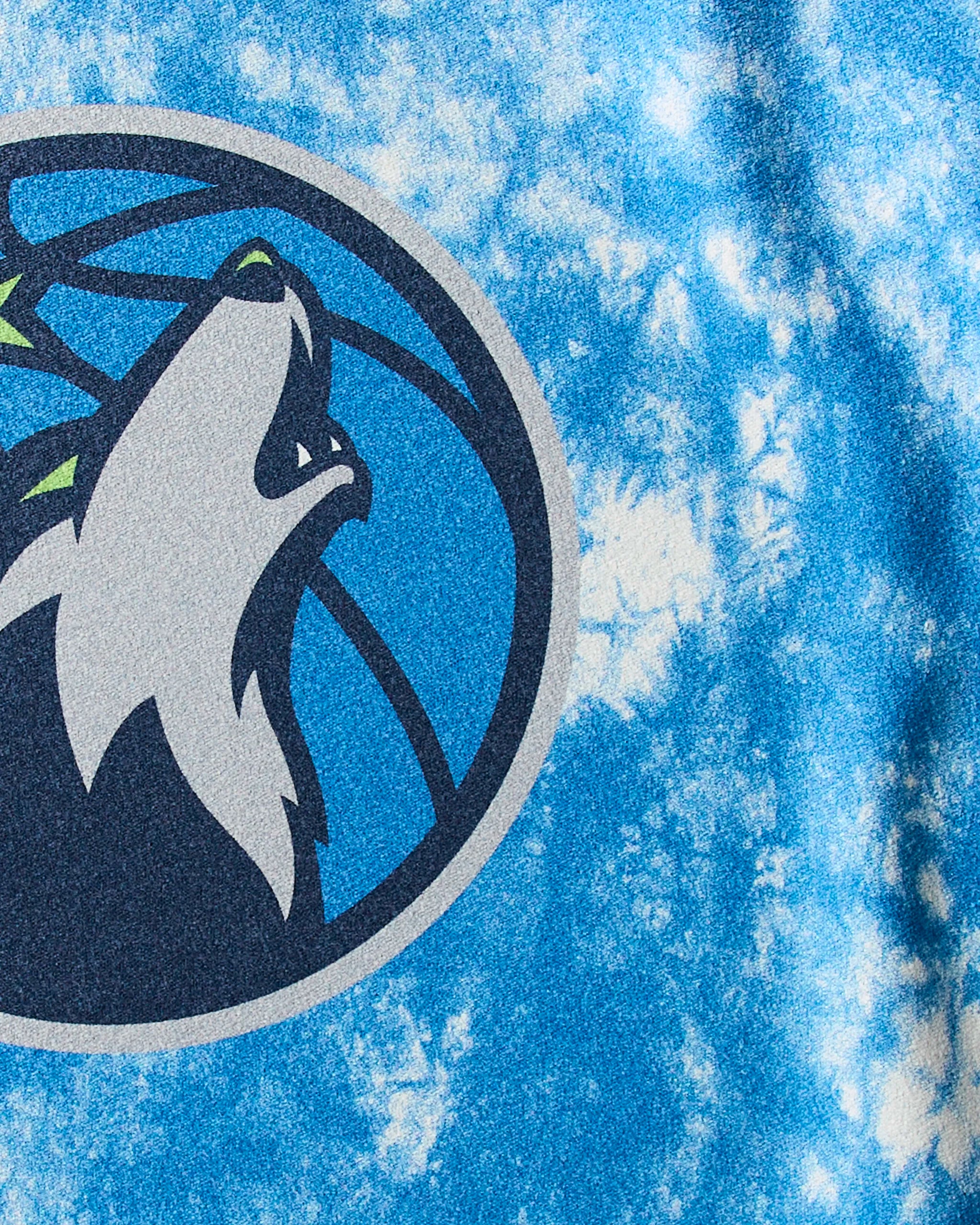 A close-up of the Timberwolves Flash logo—a stylized wolf howling before a blue basketball—appears on the Geometry NBA fitness towel with a blue and white tie-dye design.