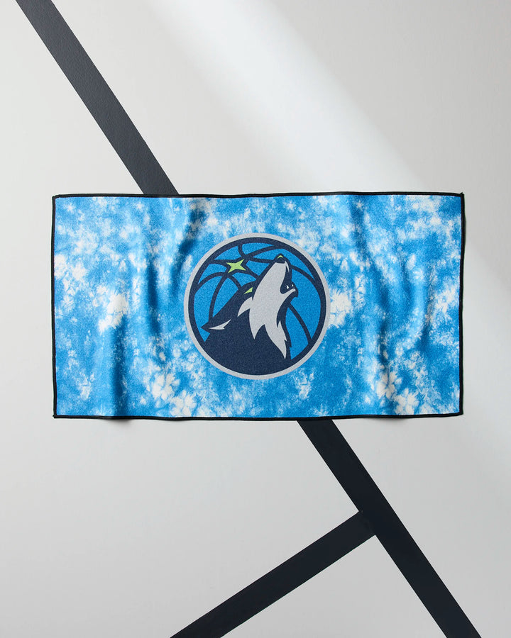 A blue and white tie-dye “Timberwolves Flash” NBA fitness towel by NBALAB x Geometry, featuring the Minnesota Timberwolves logo with a howling wolf and basketball, displayed on a white surface with black diagonal lines.
