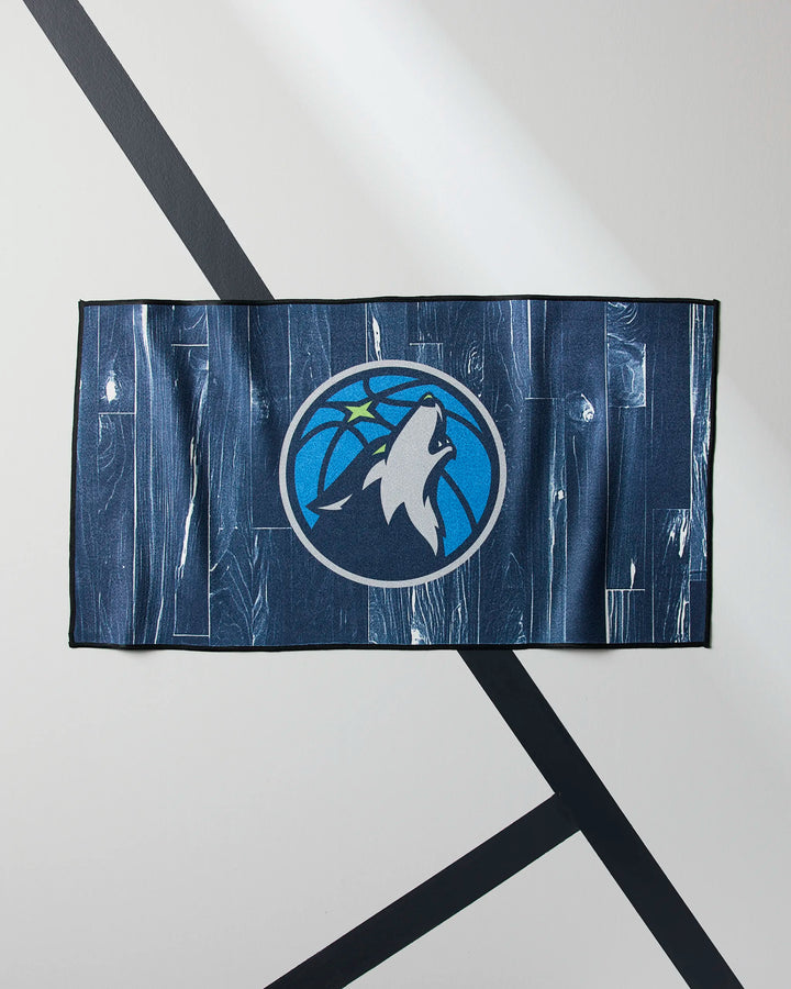 The Timberwolves Zone fitness towel by NBALAB x Geometry, in blue and featuring the Minnesota Timberwolves' howling wolf basketball logo, is shown laid flat on a light surface with black diagonal lines.