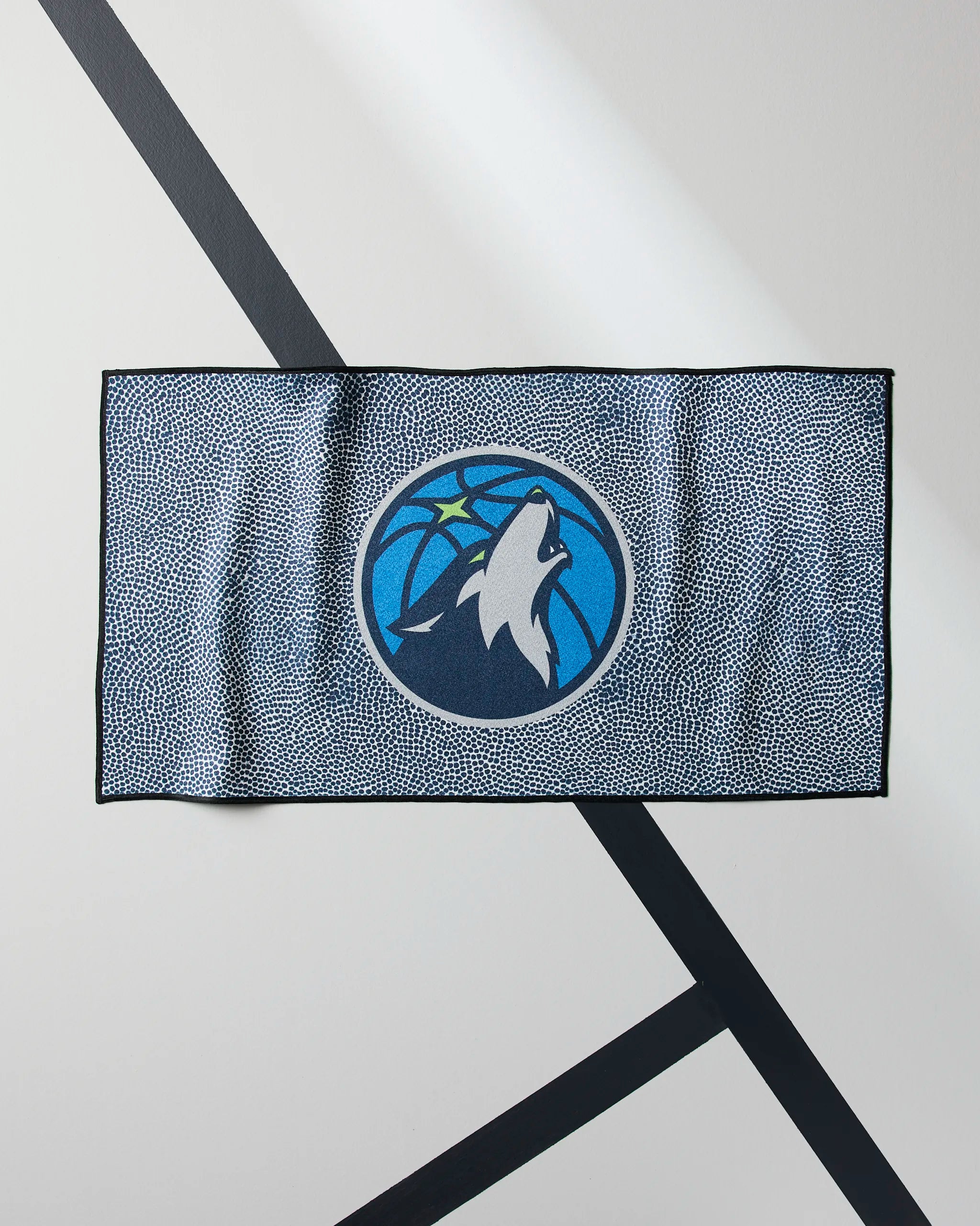 The Geometry Timberwolves Grip is a rectangular, quick-drying workout towel with a speckled blue and white design and a howling wolf basketball logo, shown on a white floor with black intersecting lines.