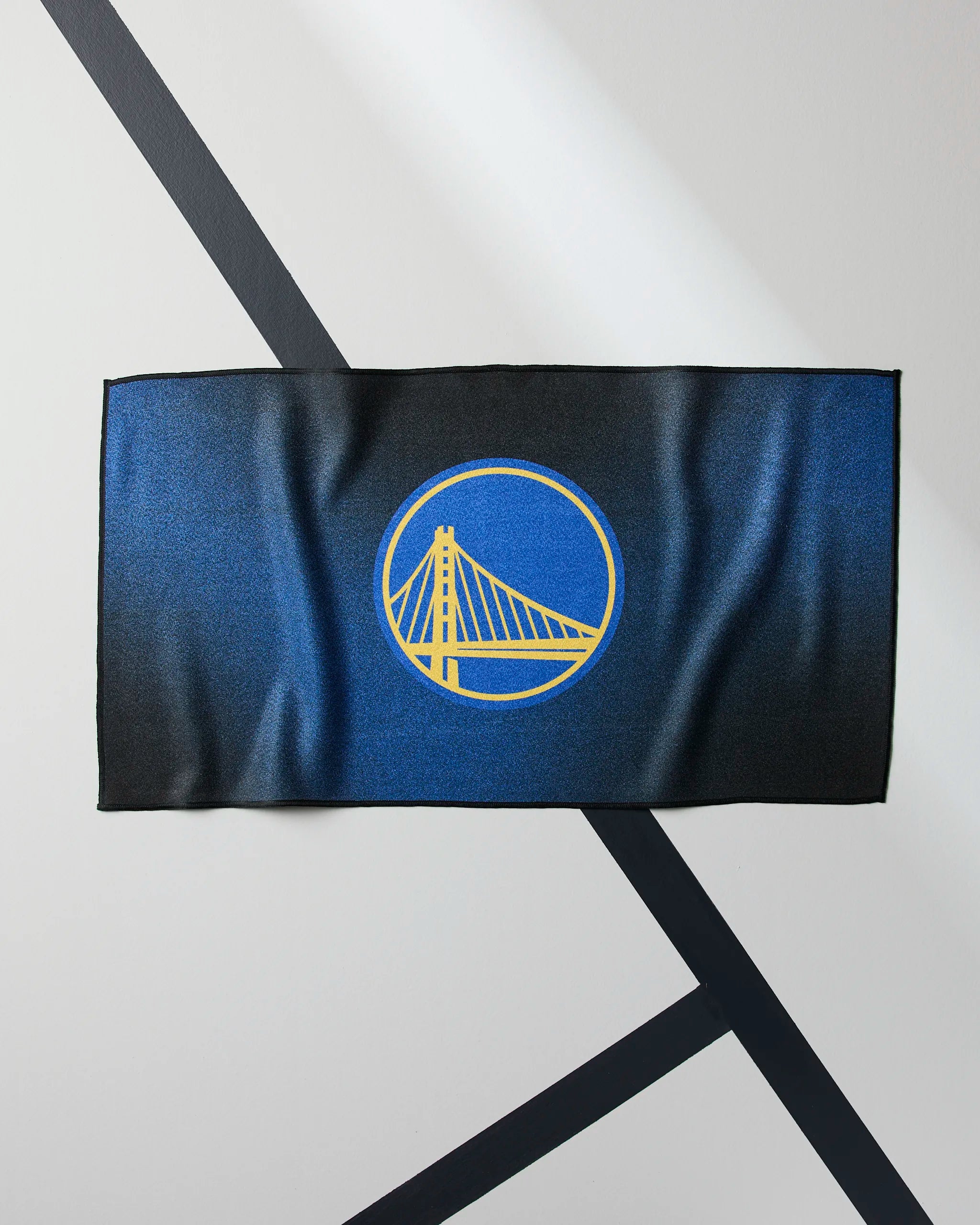 The Warriors Fade by Geometry is a rectangular NBA fitness towel featuring the Golden State Warriors bridge logo on a blue and black background, displayed on a geometric-patterned light gray surface.