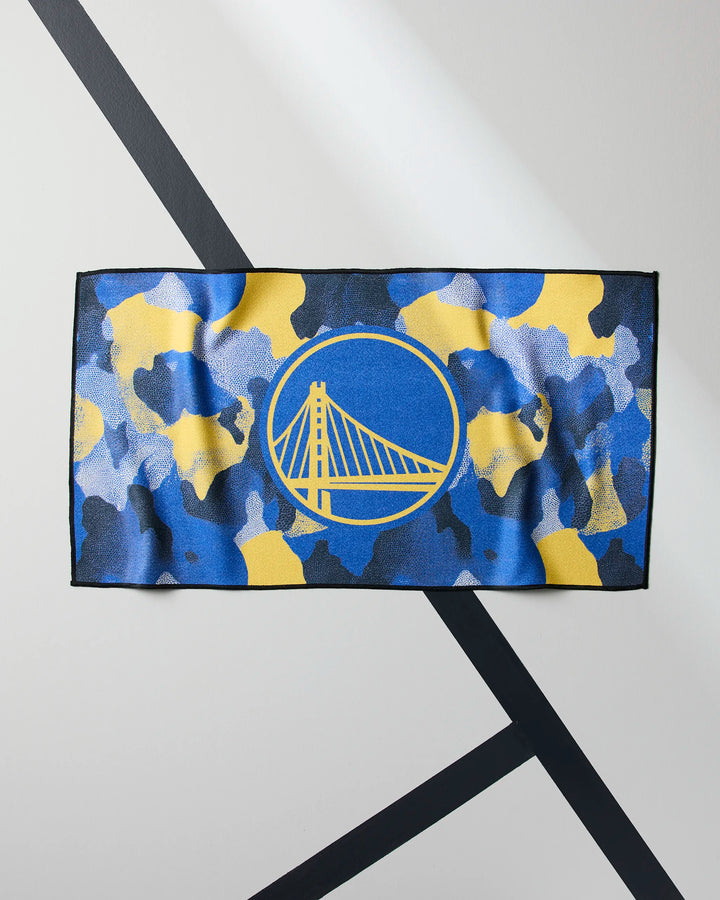 The NBALAB x Geometry "Warriors Camo" rectangular fitness towel features a blue, yellow, and black camouflage design with the Golden State Warriors logo centered, displayed on a white surface with black diagonal lines.