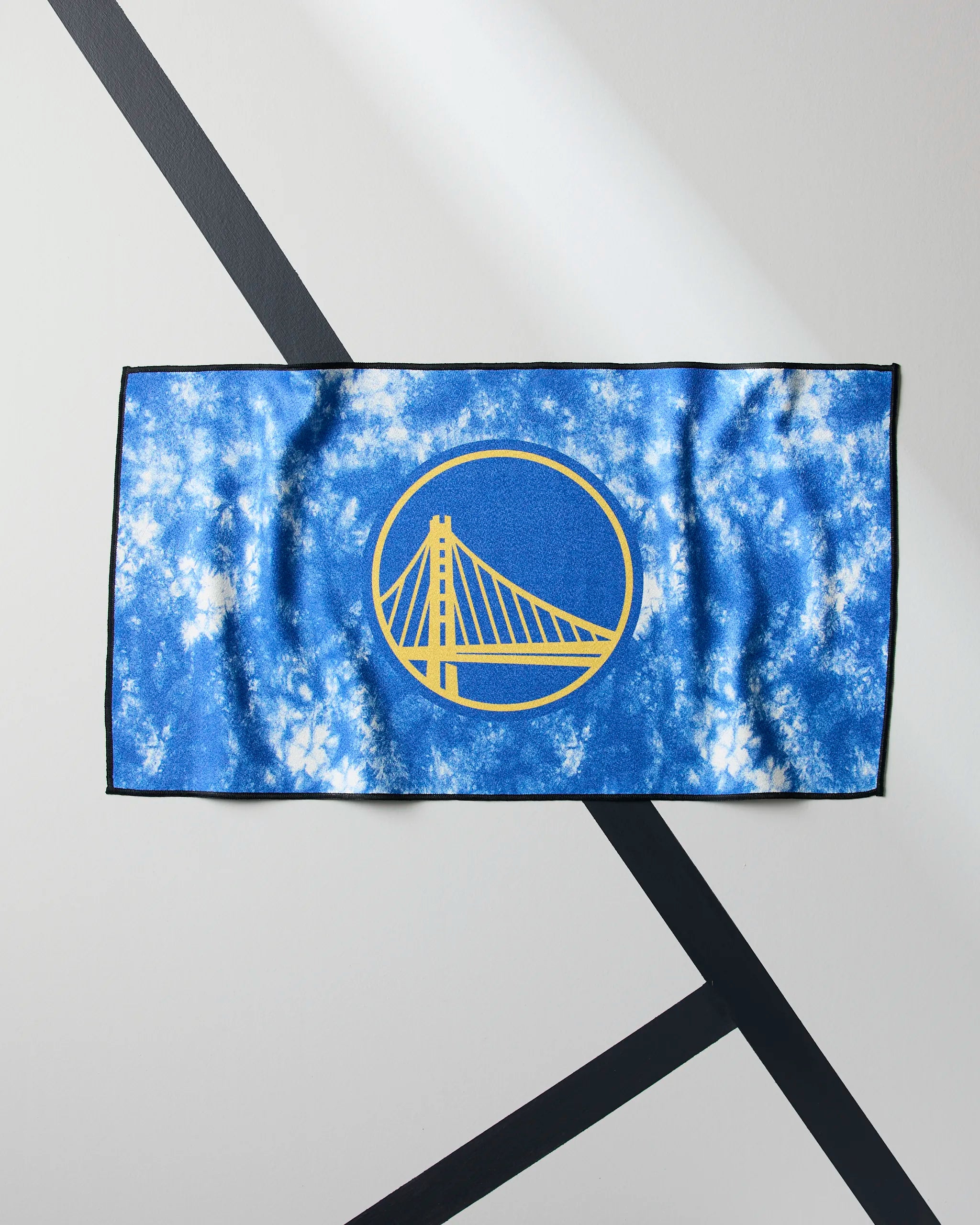 The Warriors Flash by Geometry is a blue and white tie-dye NBA fitness towel featuring the Golden State Warriors yellow bridge logo, shown flat on a light gray surface with black intersecting lines.