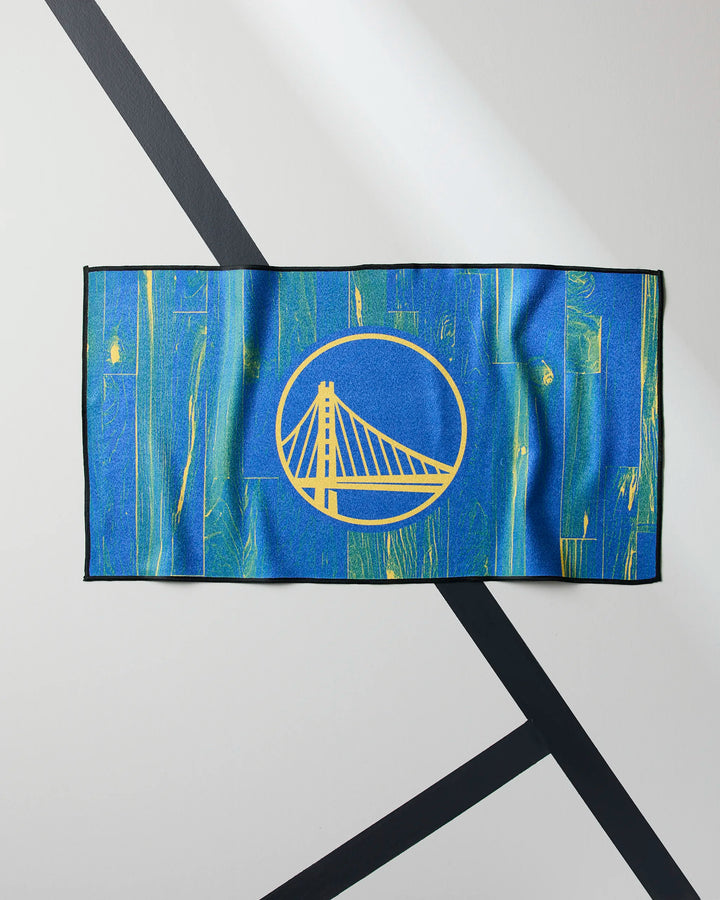 A blue Warriors Zone towel by NBALAB x Geometry, featuring the Golden State Warriors logo in yellow, hangs on a light wall with intersecting black lines.