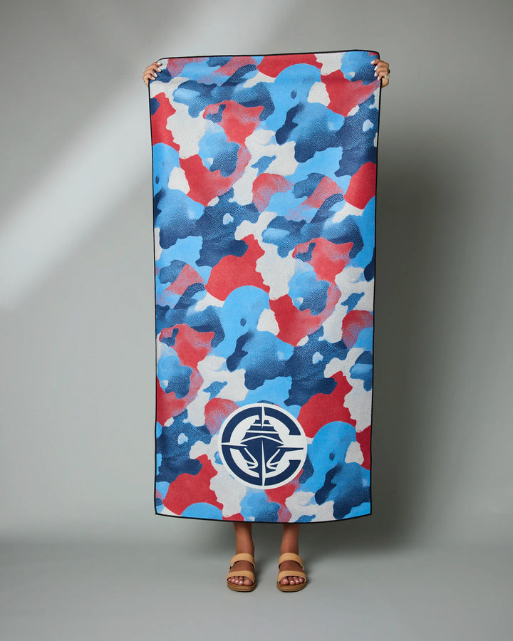 A person holds upright the NBALAB x Geometry Clippers Camo beach towel featuring a blue, red, and white camo pattern and a large circular compass logo; only their hands and sandaled feet can be seen.