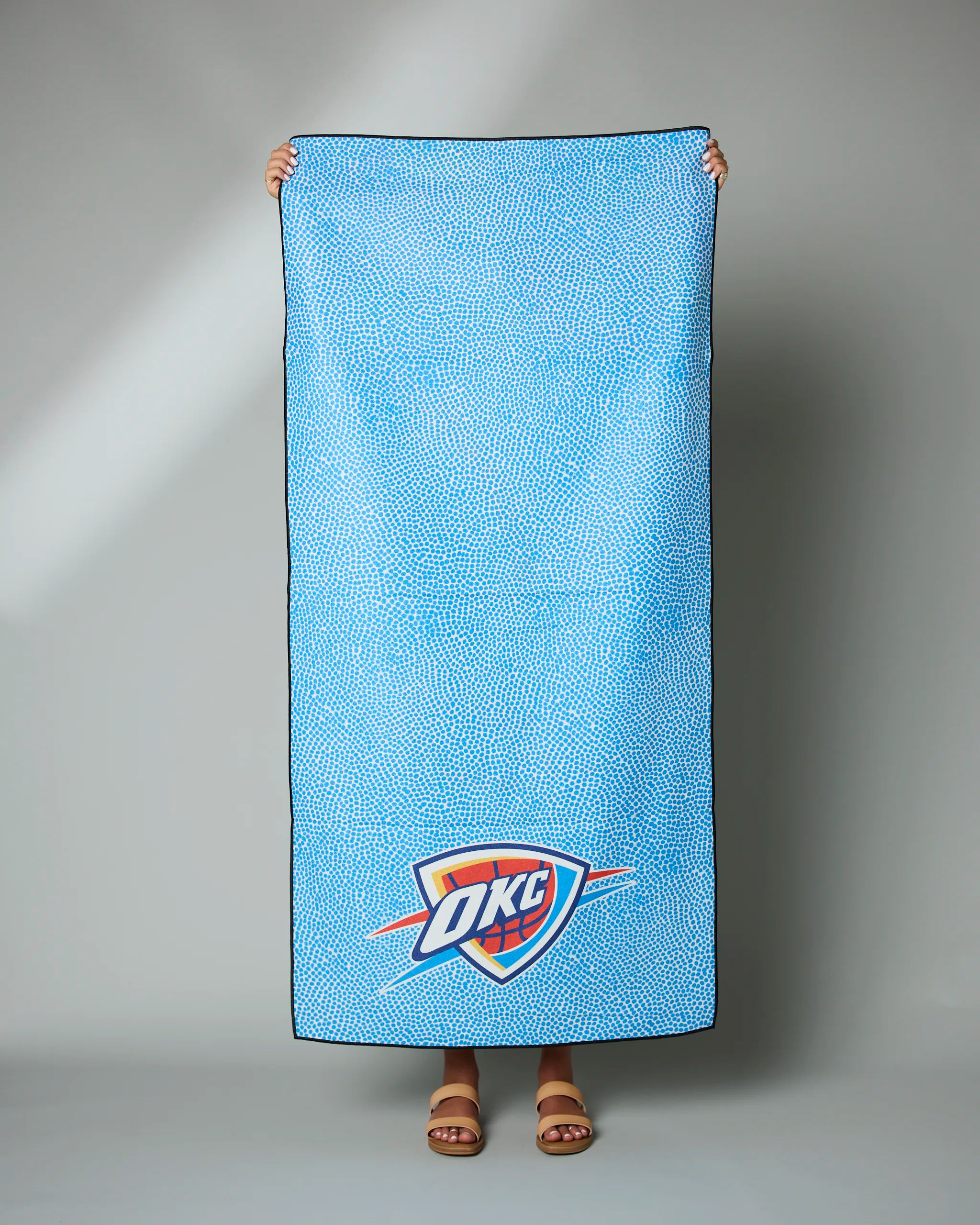 A person holds up the Geometry OKC Grip, a blue rectangular towel with a dotted pattern and the OKC Thunder logo near the bottom; only their hands and feet in sandals are visible.