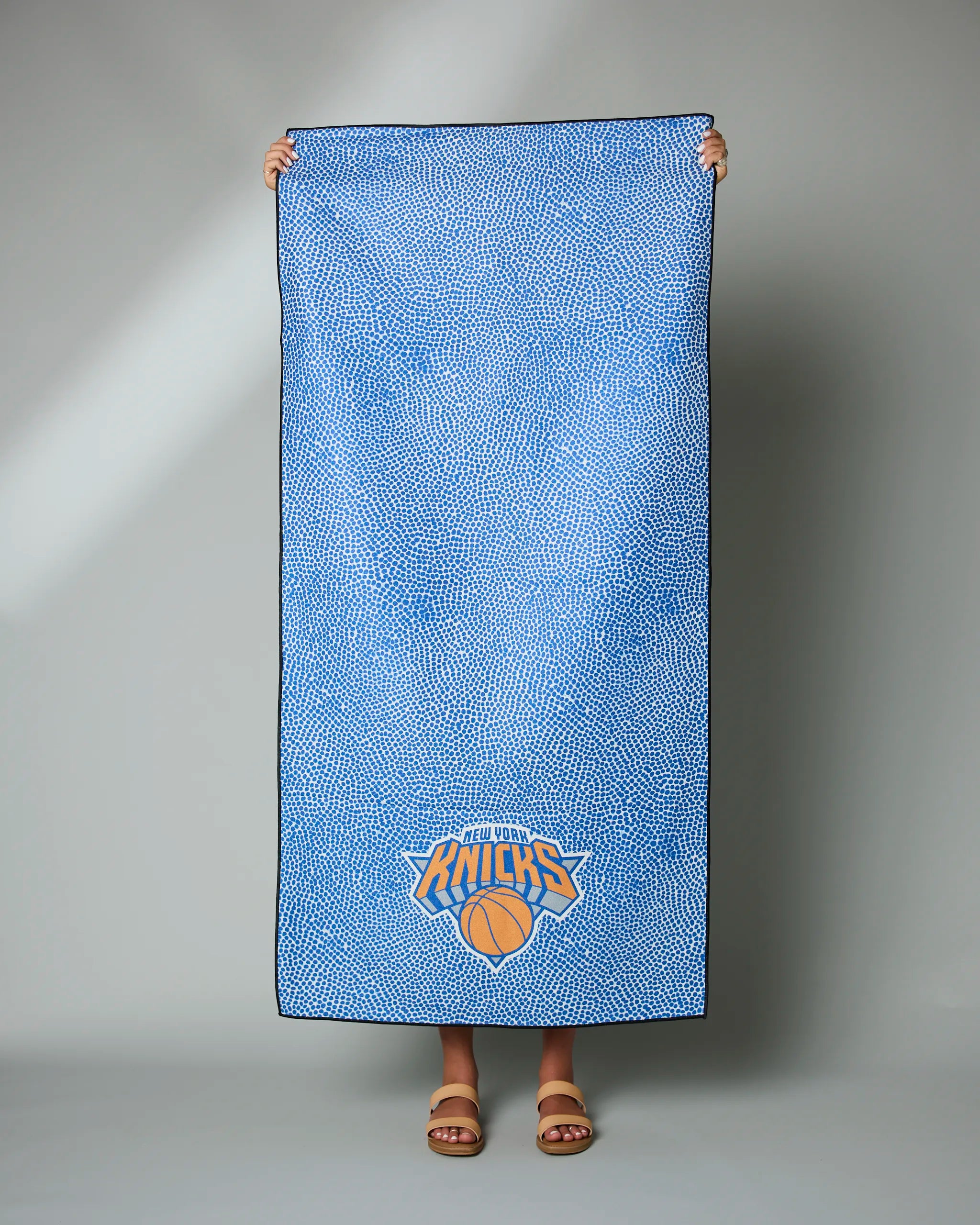 A person holds up the Geometry Knicks Grip, a large blue towel with a white speckled pattern and the New York Knicks logo near the bottom; only their hands and sandaled feet are visible.