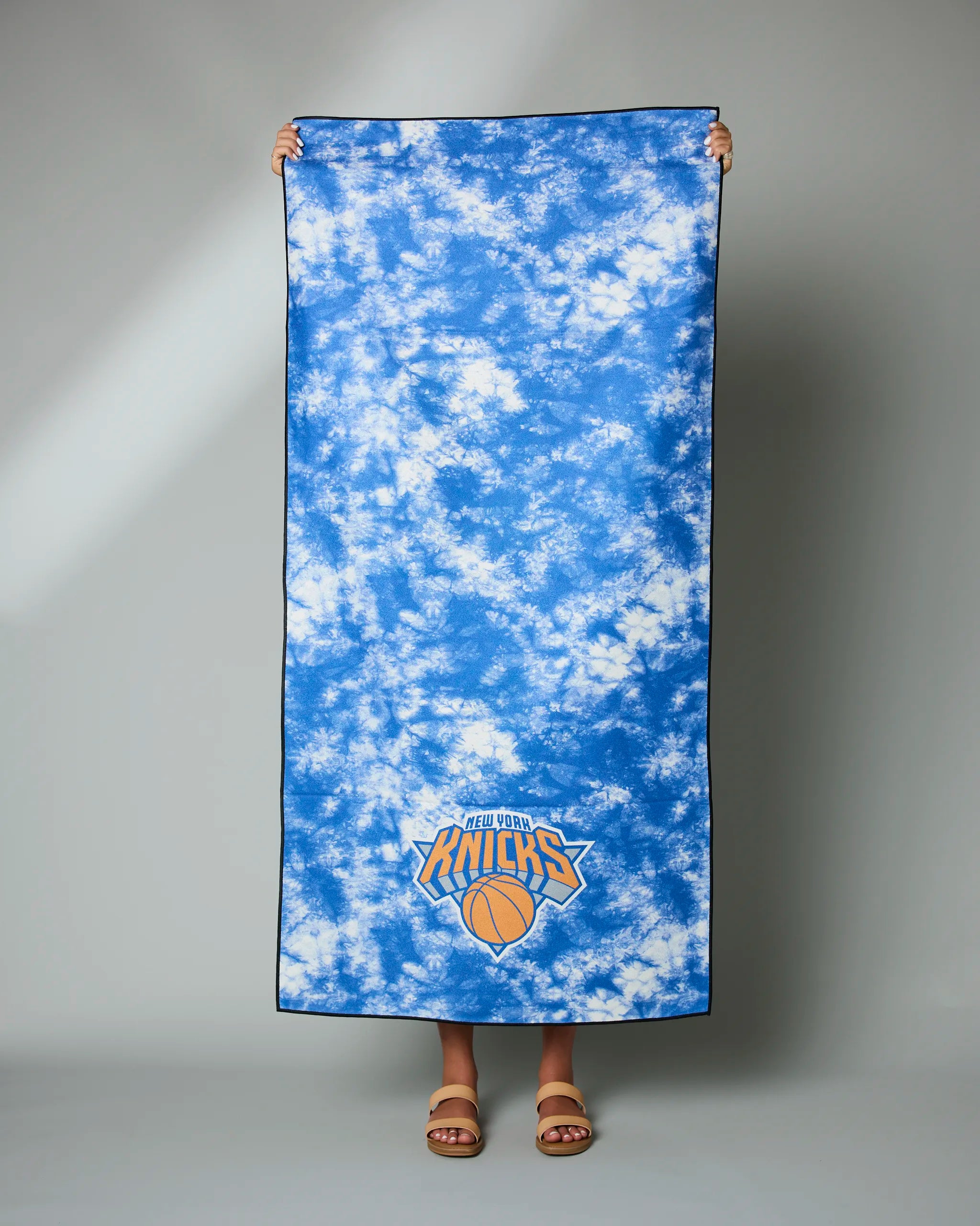A person stands holding the Knicks Flash towel by Geometry—a large blue and white tie-dye towel featuring the New York Knicks logo at the bottom, with only their feet in sandals visible.