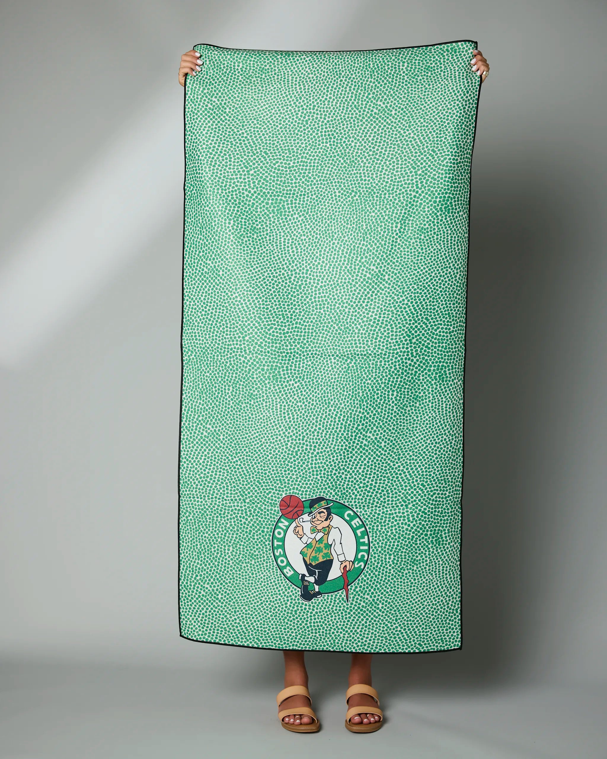 A person stands barefoot in sandals against a plain background, holding the Geometry Celtics Grip towel—a green towel with small white dots and the Boston Celtics logo near the bottom.