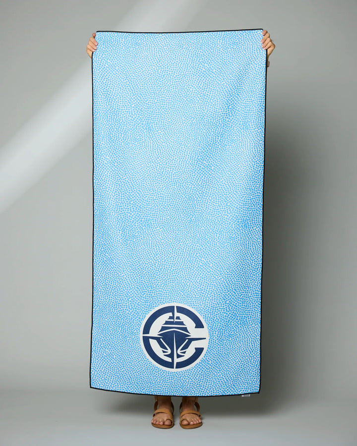 A person holds up the NBALAB x Geometry Clippers Grip, a light blue recycled beach towel with a subtle dotted pattern and a white nautical logo near the bottom; only their feet and hands are visible.