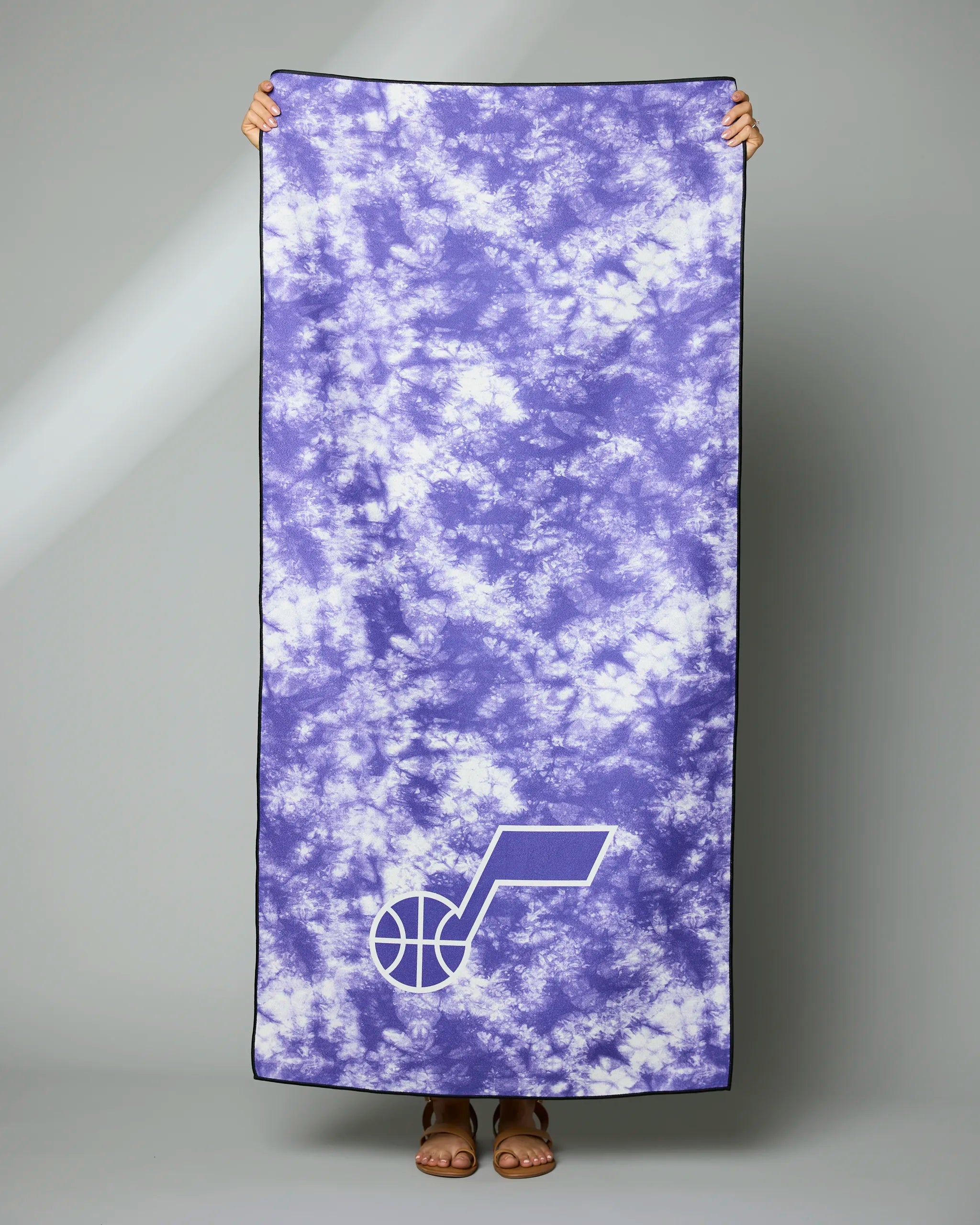 A person holds the Jazz Flash towel by Geometry, featuring a purple and white tie-dye design and the Utah Jazz logo in white at the bottom right corner. The persons head is not visible, shown against a plain background.