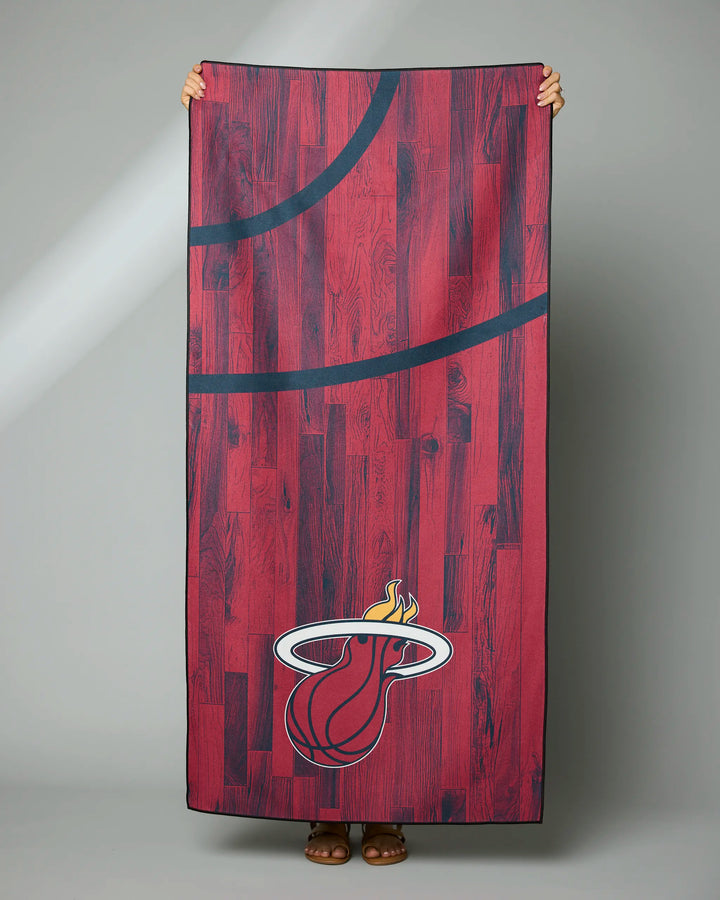 A person holds up the Heat Zone NBA beach towel by NBALAB x Geometry, featuring a red wood floor pattern with black court lines and the Miami Heat flaming basketball logo at the bottom.