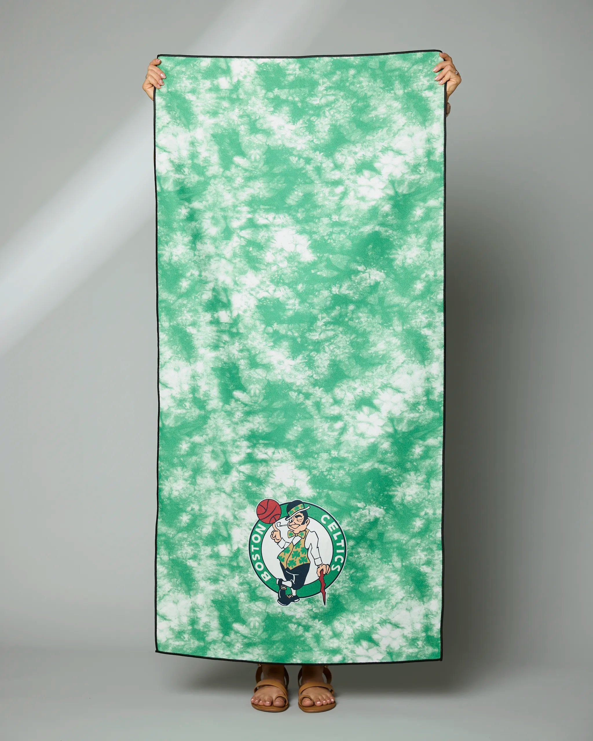 A person holds the Geometry Celtics Flash towel, featuring a large green-and-white tie-dye design with the Boston Celtics logo at the bottom; only their hands and feet are visible.