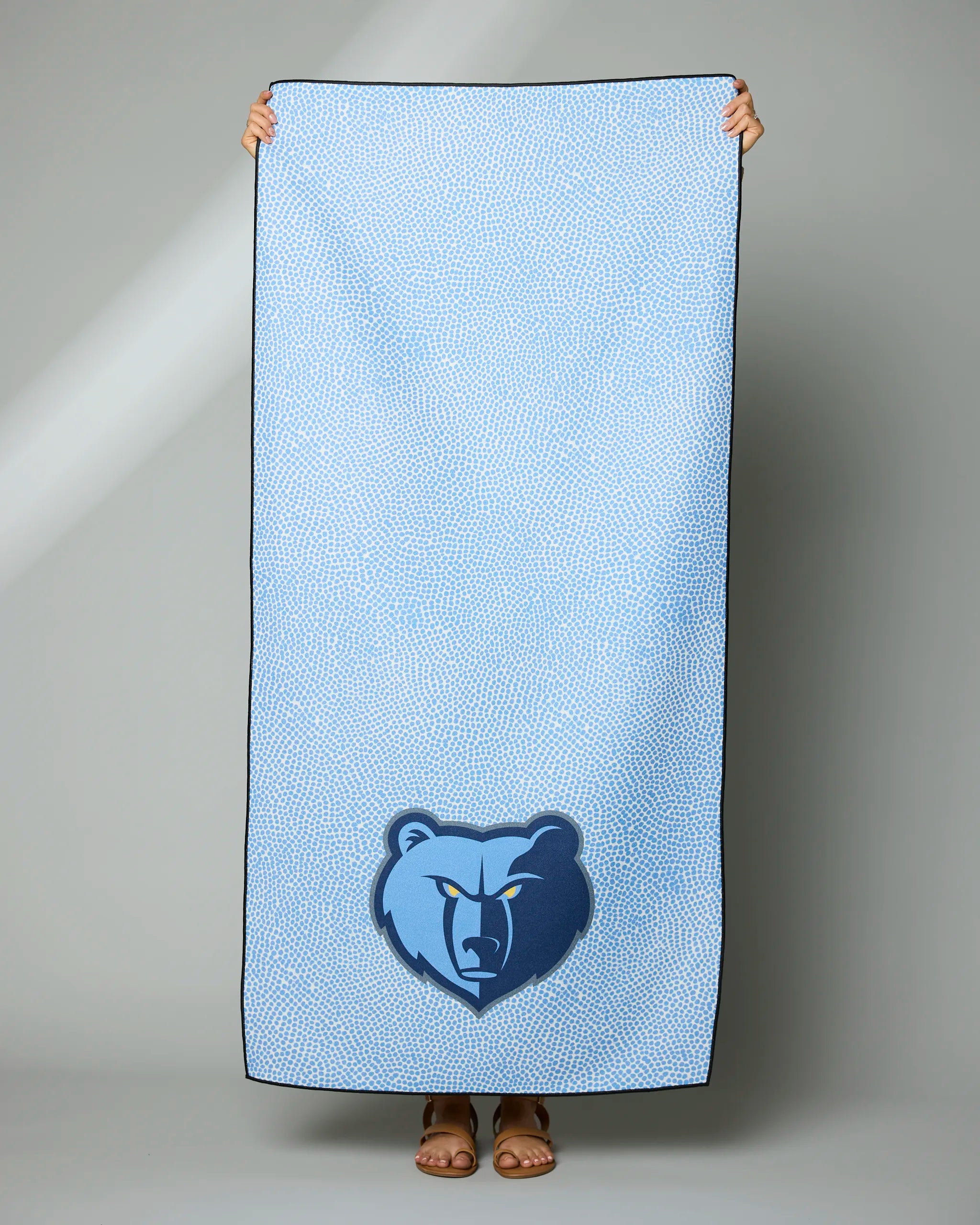A person holds up the Geometry Grizzlies Grip towel, featuring a light blue design with the Memphis Grizzlies bear logo near the bottom. Only their hands and feet are visible; the rest of their body is hidden behind the towel.