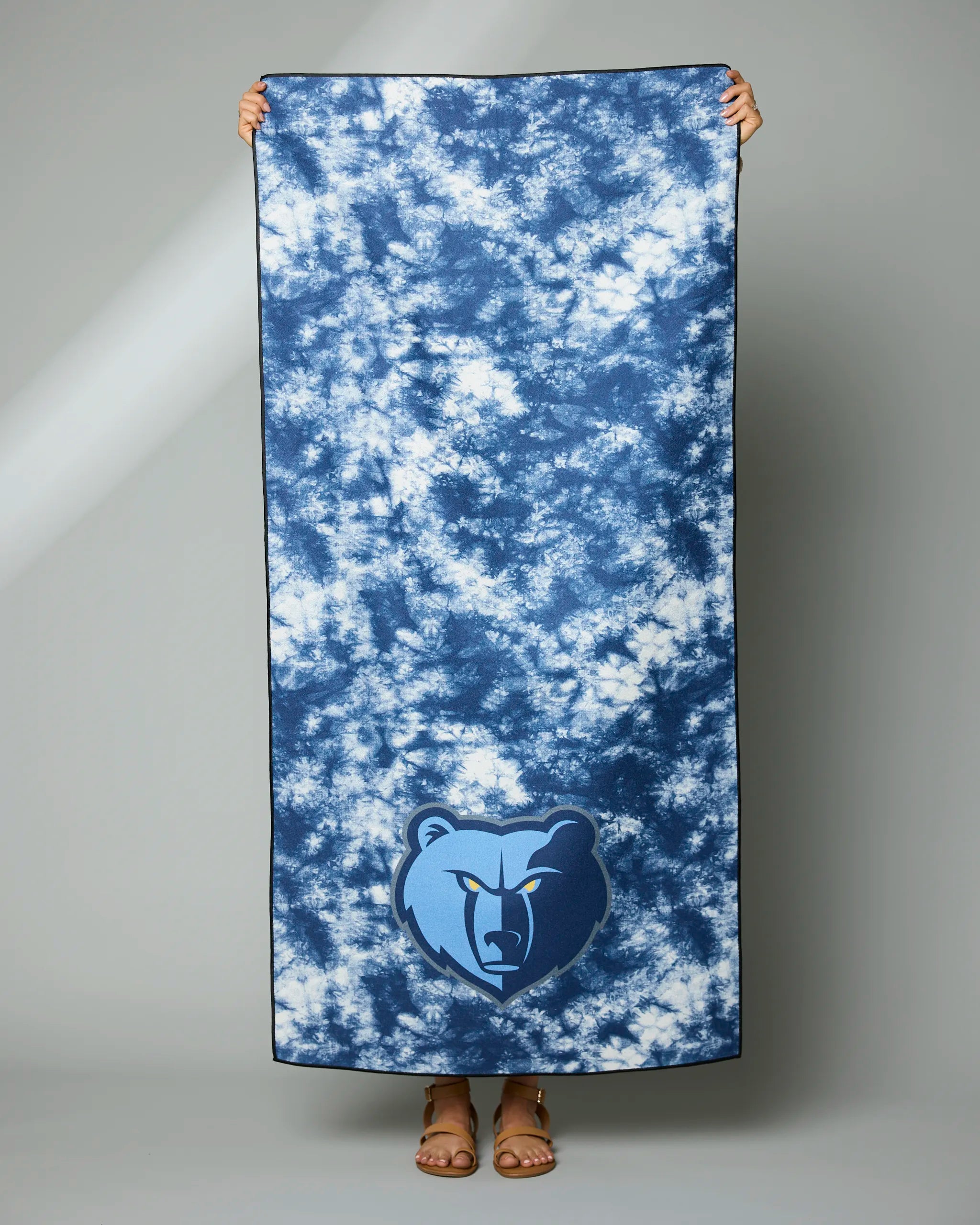A person holds up the Geometry Grizzlies Flash towel, featuring the Memphis Grizzlies bear logo at the bottom. Their face is hidden behind the large blue tie-dye towel, with only their hands and feet visible.