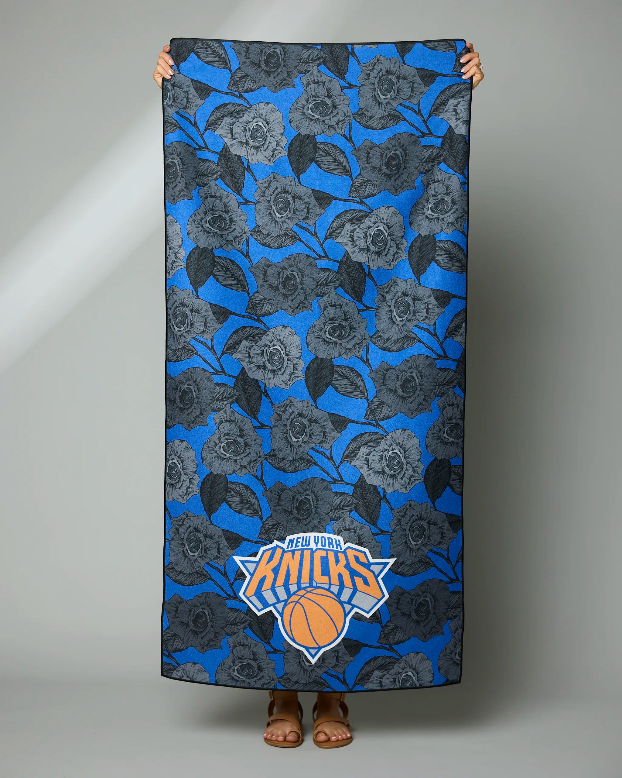 A person displays the Geometry Knicks Floral towel, featuring a blue and gray floral design with the New York Knicks logo at the bottom. Only their hands and feet are visible.